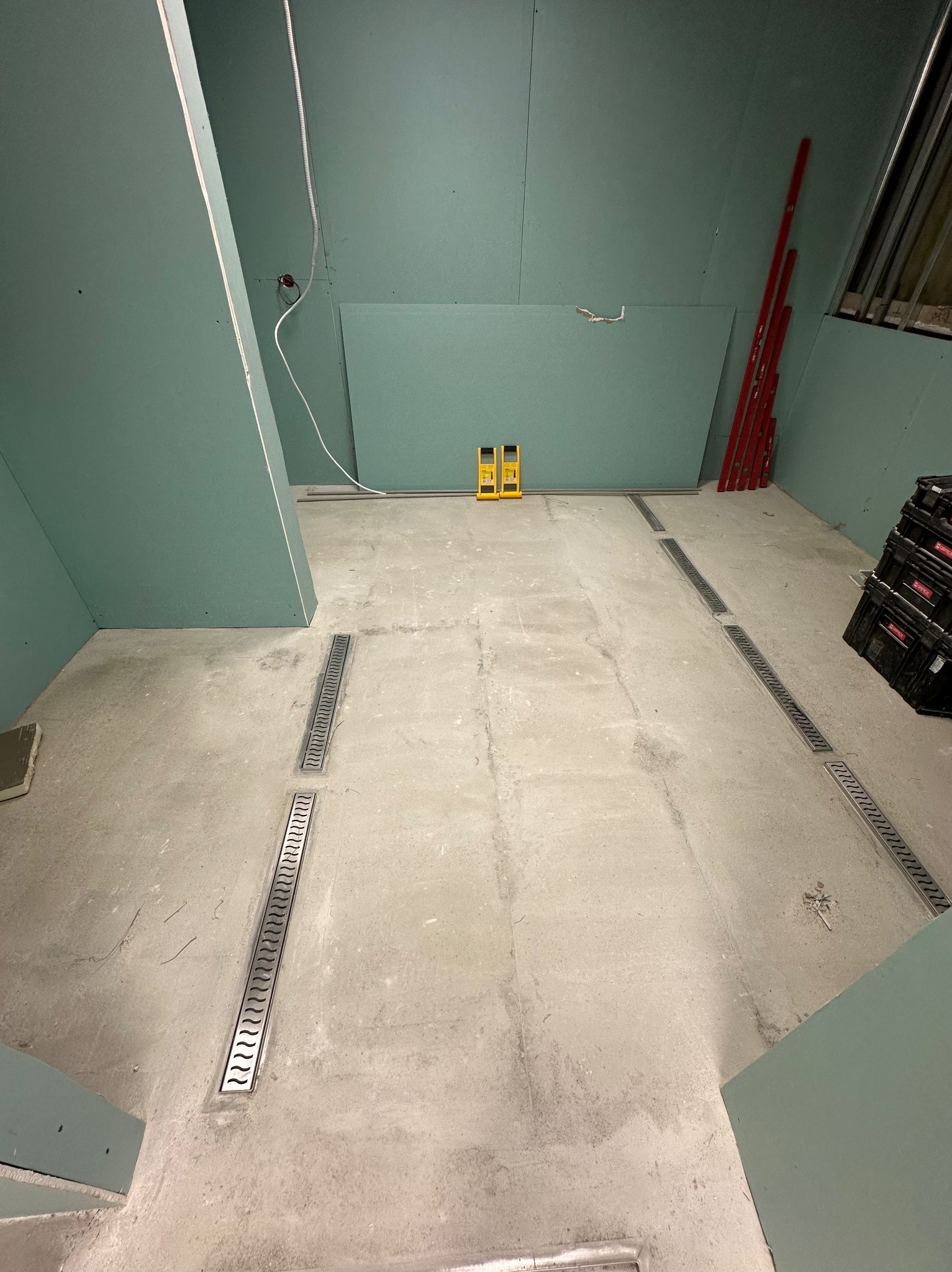 Surowa łazienka w trakcie prac: zielone płyty GK na ścianach, w posadzce liniowe odpływy. Widoczne poziomice i narzędzia. Etap przygotowania do dalszych prac wykończeniowych.