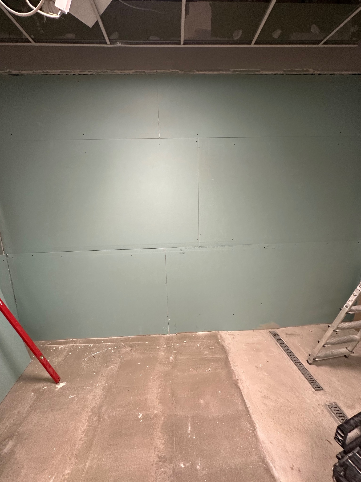 Ściana pokryta płytami kartonowo-gipsowymi, widoczne łączenia. Na podłodze resztki materiałów budowlanych, drabina oparta o ścianę. Sufit podwieszany w tle.