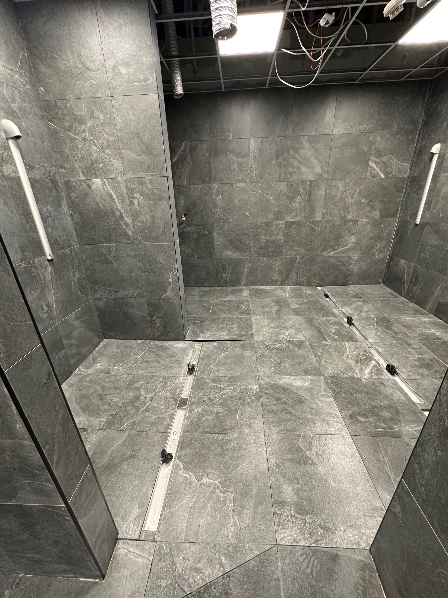 Wykończona łazienka z szarymi płytkami imitującymi kamień, z odpływem liniowym w podłodze. Widoczne fragmenty instalacji pod sufitem.