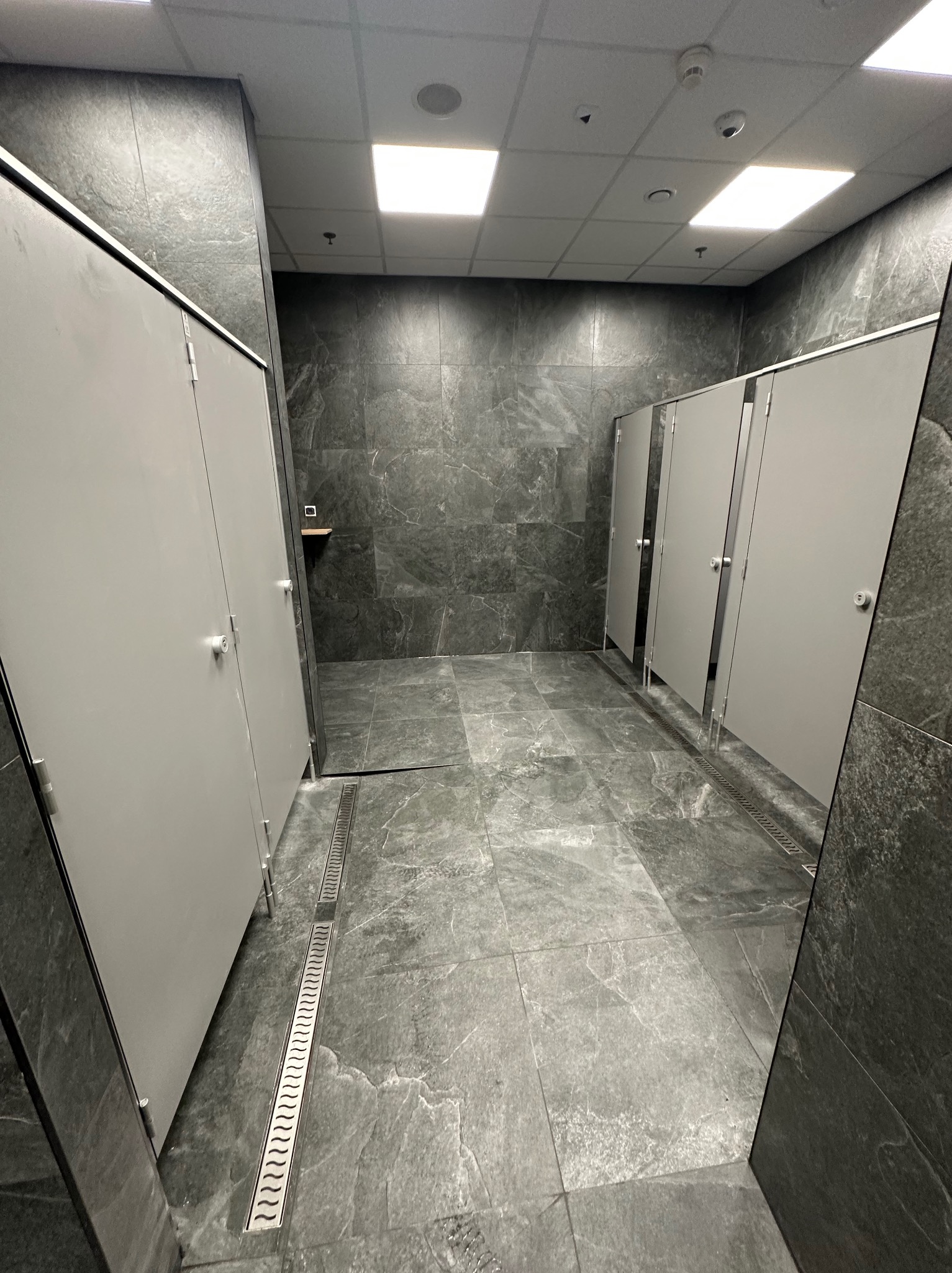 Nowoczesna toaleta publiczna z szarymi płytkami imitującymi kamień i kabinami z jasnymi drzwiami. Widoczne odpływy liniowe w podłodze.