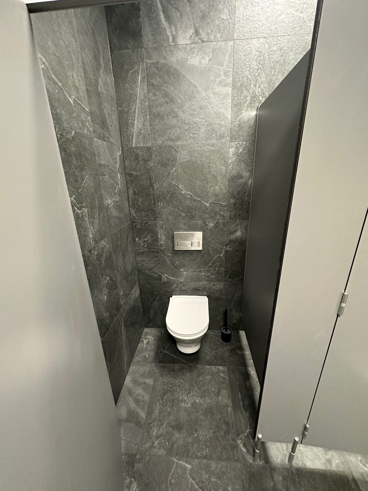 Nowoczesna toaleta z szarymi płytkami imitującymi kamień, wiszącą muszlą i minimalistycznym przyciskiem spłukującym. Widok z góry, podkreślający geometrię i fakturę materiałów.