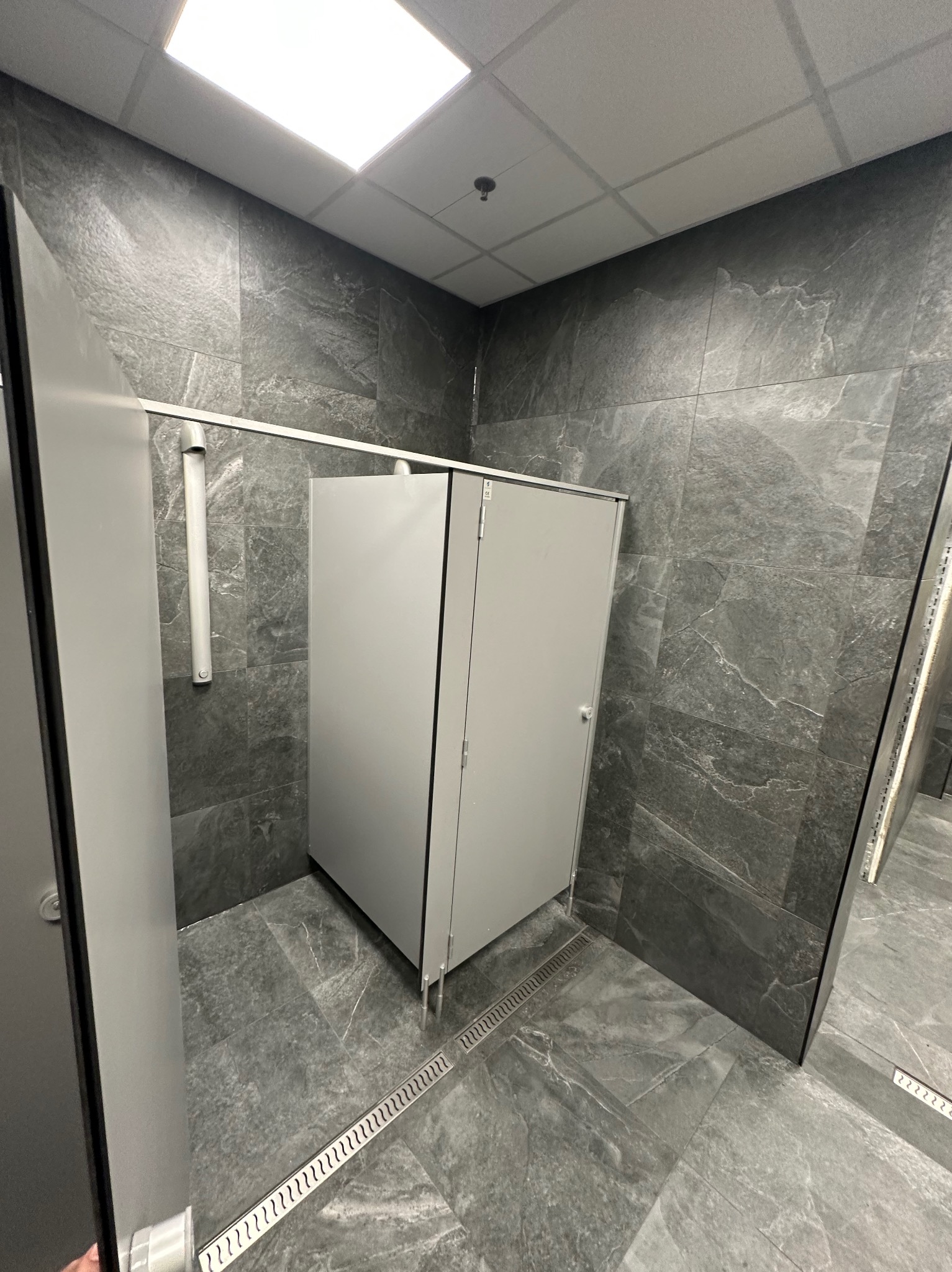 Nowoczesna toaleta publiczna z szarymi płytkami imitującymi kamień na ścianach i podłodze. Kabiny WC z metalowymi elementami i odpływem liniowym w podłodze.