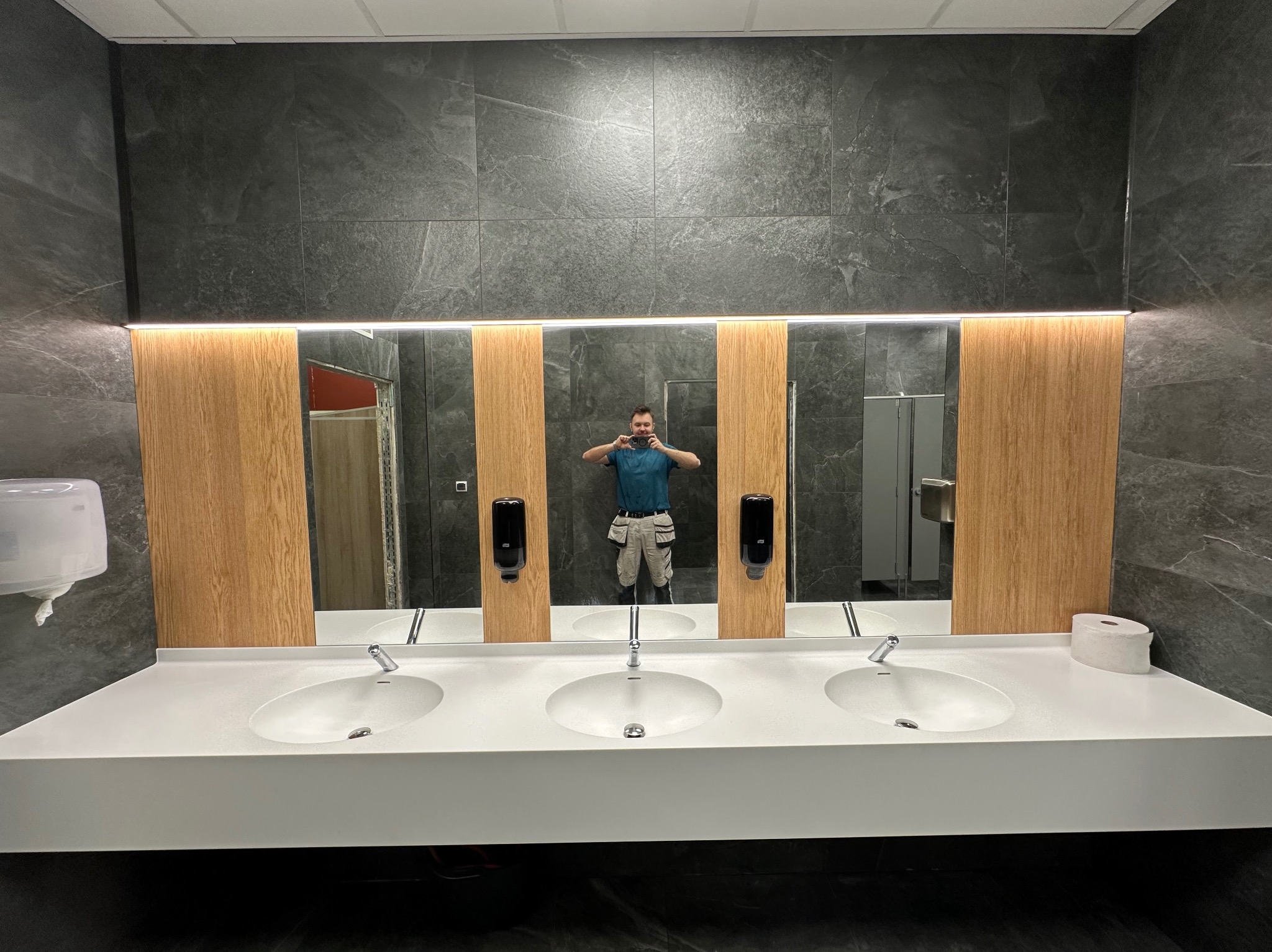 Nowoczesna łazienka publiczna z trzema umywalkami, szarą okładziną ścienną i drewnianymi panelami. Osoba robiąca zdjęcie widoczna w lustrze.