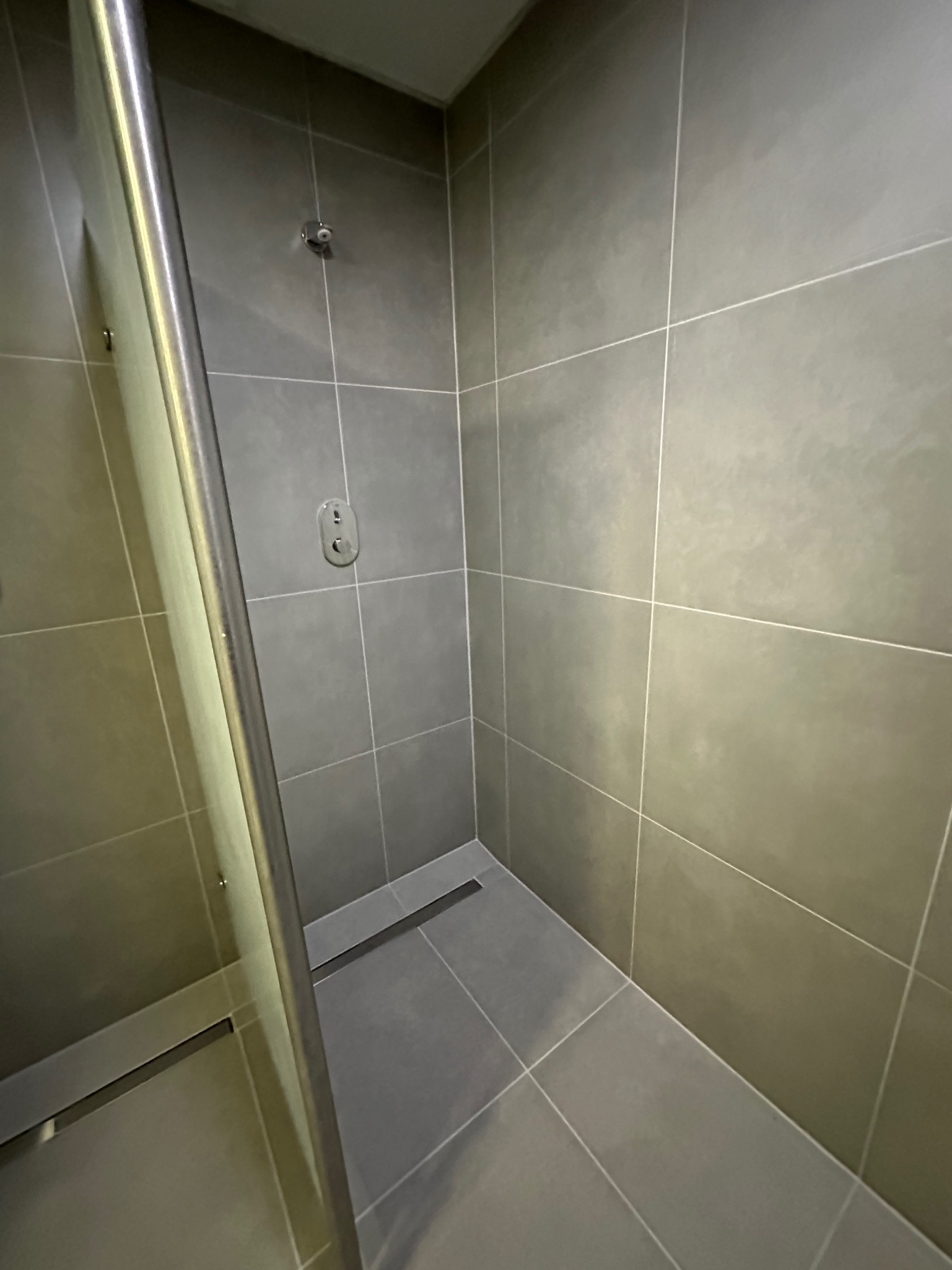 Nowoczesna kabina prysznicowa z szarymi, kwadratowymi płytkami, odpływem liniowym i minimalistyczną armaturą. Widok z perspektywy wejścia, z delikatnym oświetleniem.