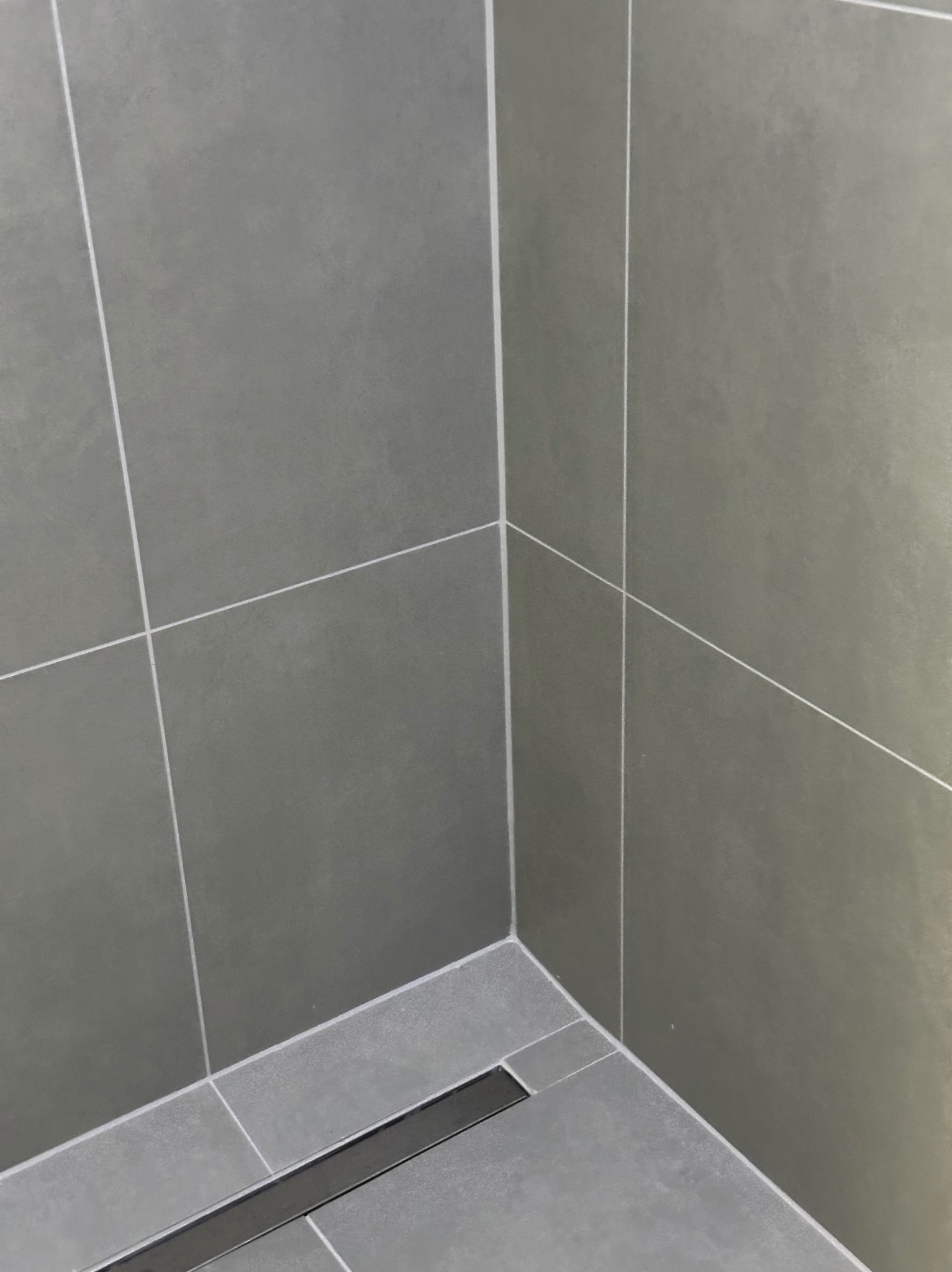 Szare, matowe płytki w kabinie prysznicowej z odpływem liniowym. Minimalistyczny design, fuga w kolorze białym. Ujęcie z narożnika, widoczny fragment podłogi i ścian.