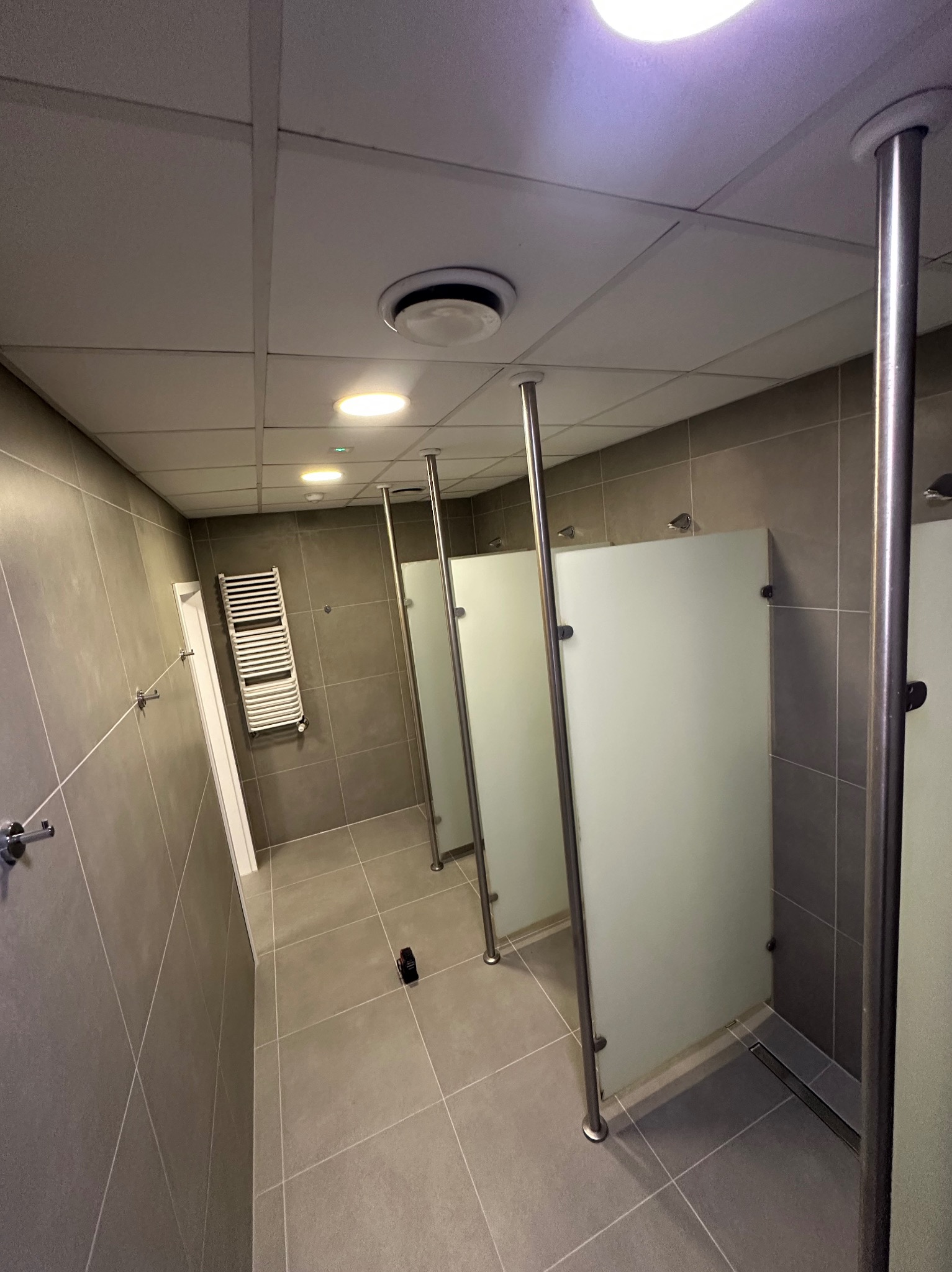 Nowoczesna łazienka publiczna z kabinami WC o matowych szklanych ściankach i metalowych stelażach, szare płytki na ścianach i podłodze, oświetlenie sufitowe.