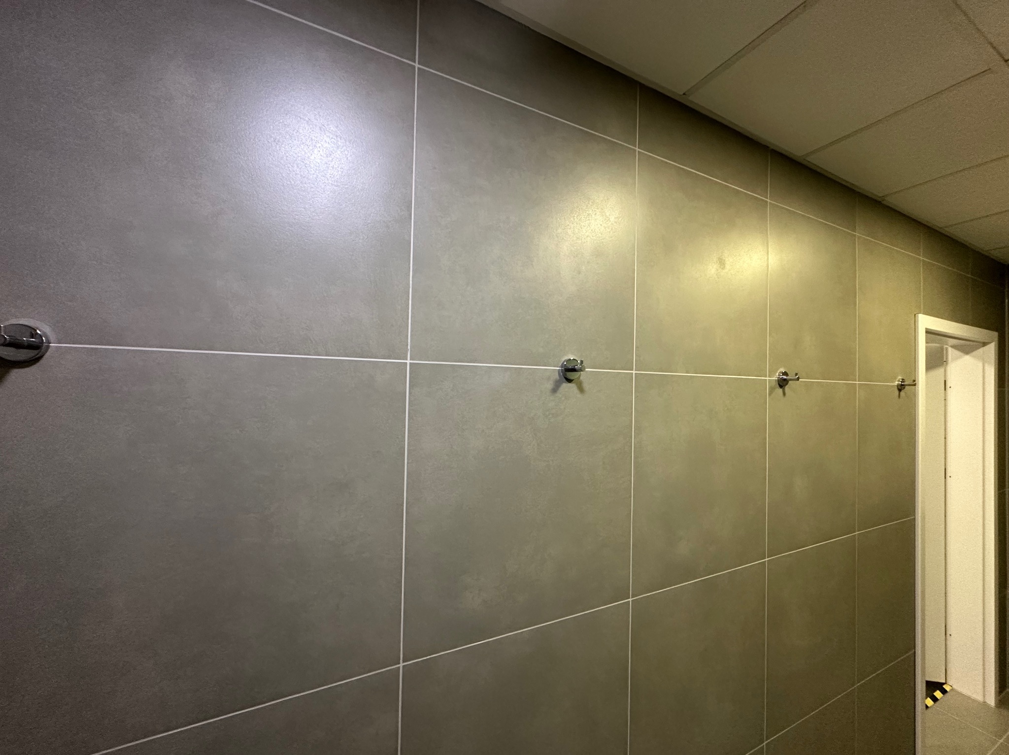 Szare, matowe płytki ścienne z fugami w jasnym odcieniu, rozmieszczone równomiernie. Na ścianie widoczne metalowe wieszaki. Ujęcie z perspektywy, widoczny fragment sufitu i drzwi.