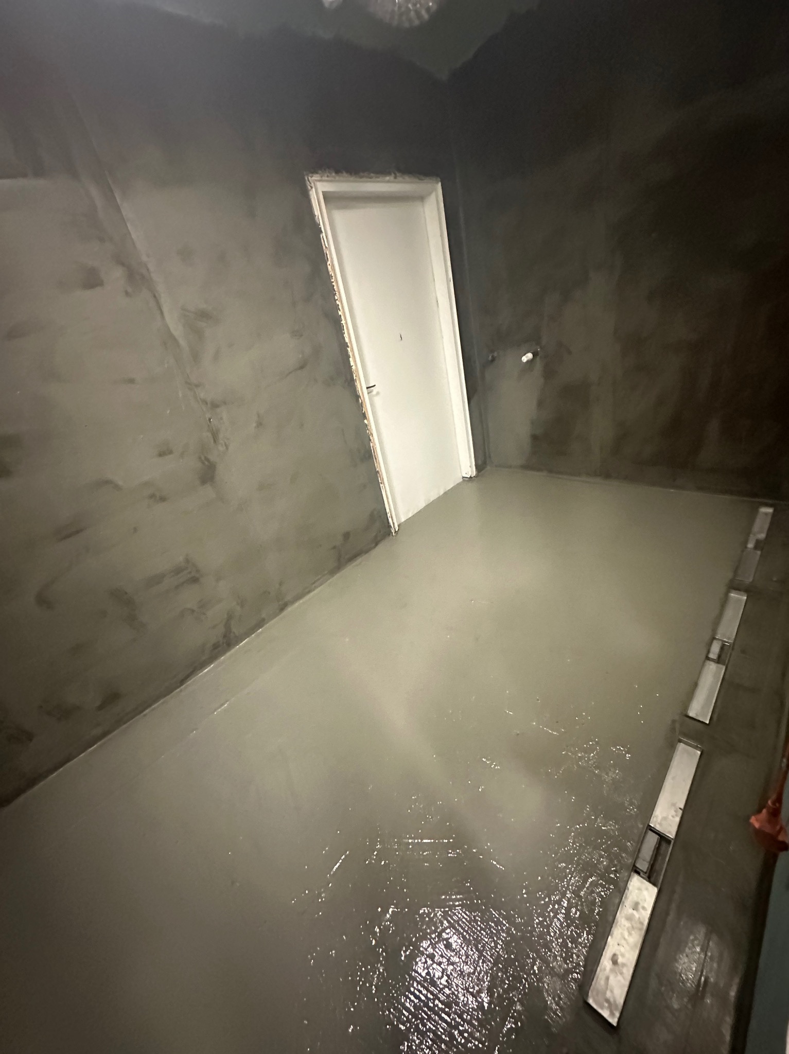 Wnętrze łazienki w trakcie prac: ściany pokryte szarą warstwą, świeżo wylana, mokra posadzka z widocznymi odpływami liniowymi. Białe drzwi w tle.