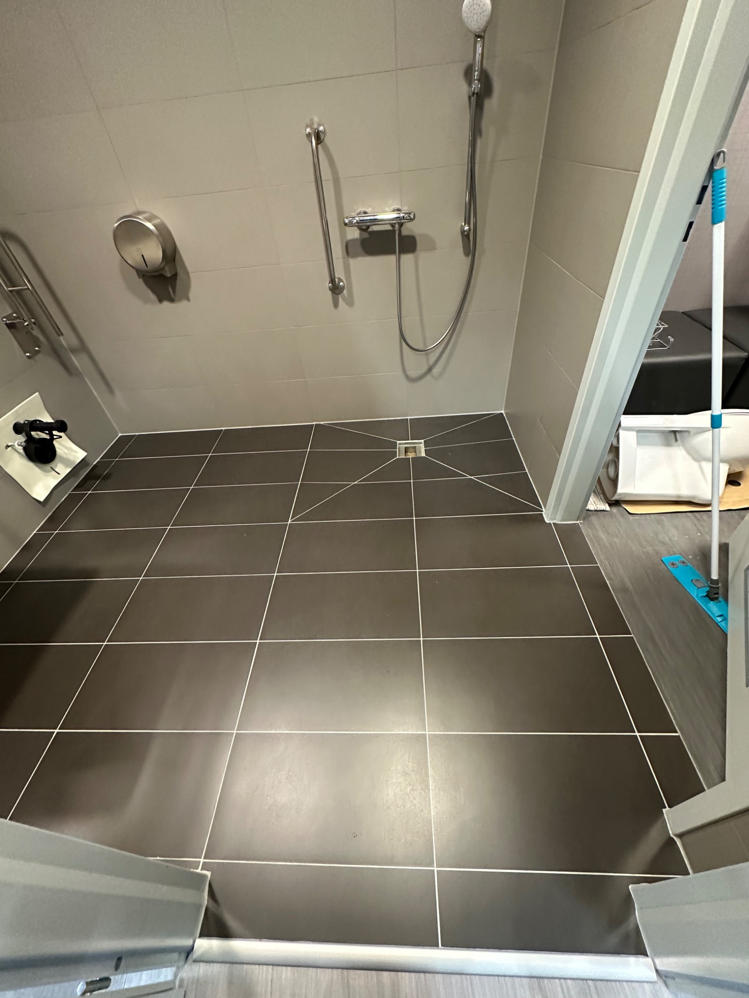 Nowoczesna kabina prysznicowa z ciemnymi płytkami podłogowymi i jasnymi fugami, wyposażona w uchwyty dla niepełnosprawnych i odpływ liniowy.