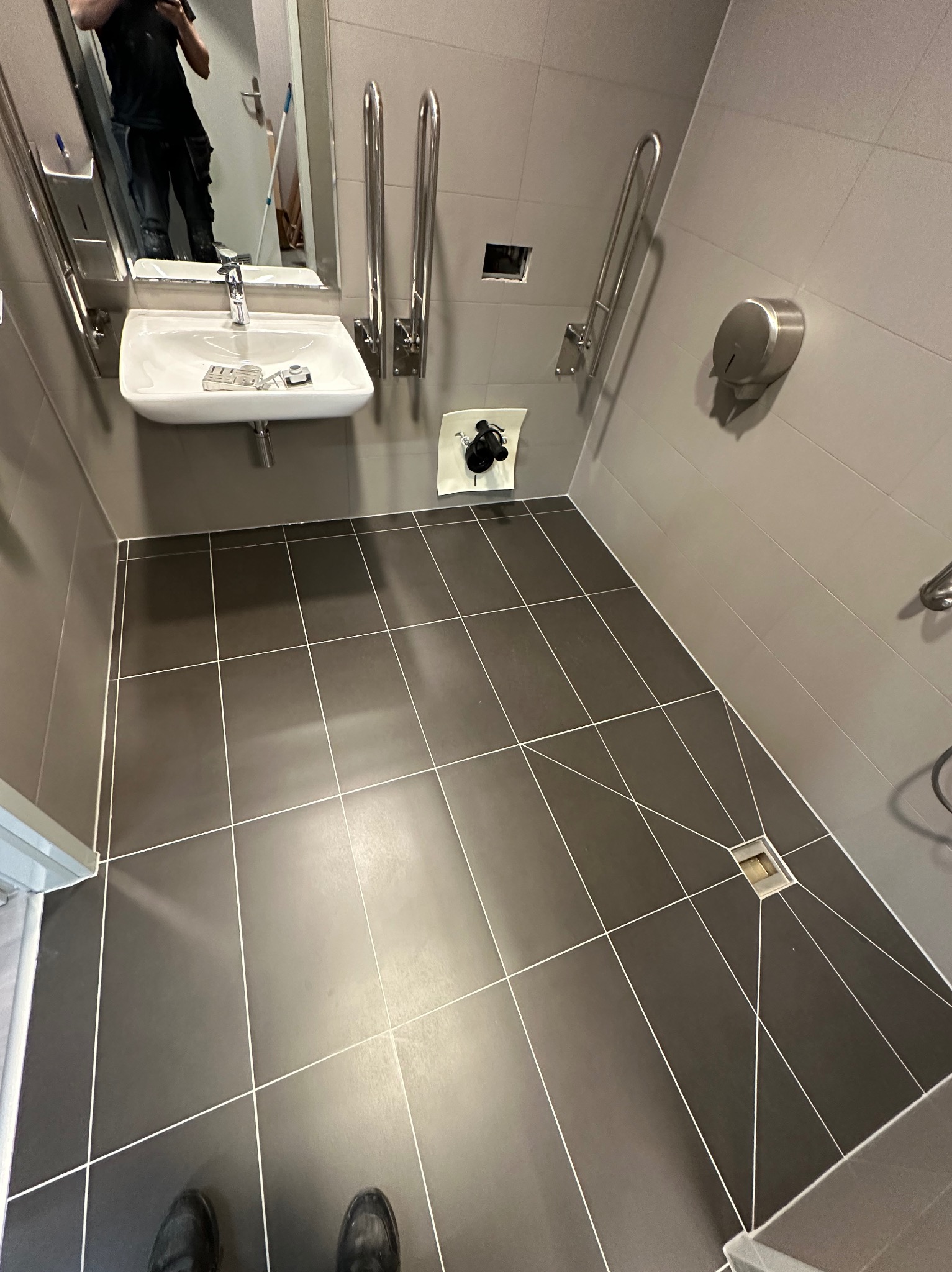 Wykończona łazienka w szarościach z umywalką i uchwytami dla niepełnosprawnych. Ciemne płytki podłogowe z jasną fugą, odpływ liniowy. Widoczne odbicie osoby w lustrze.