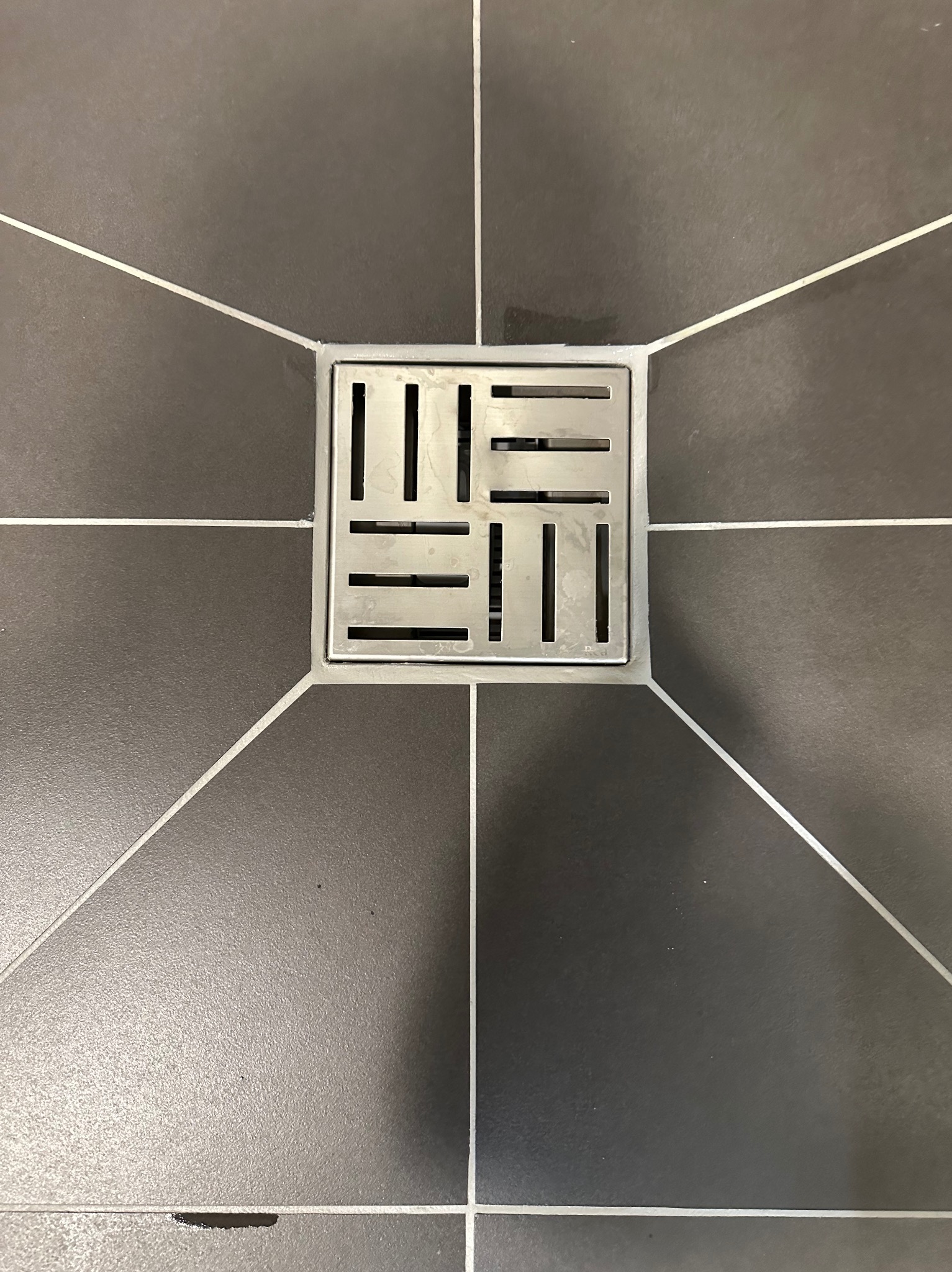 Ciemne, kwadratowe płytki podłogowe z fugami w kształcie gwiazdy, z centralnie umieszczonym, metalowym odpływem liniowym. Minimalistyczny design.