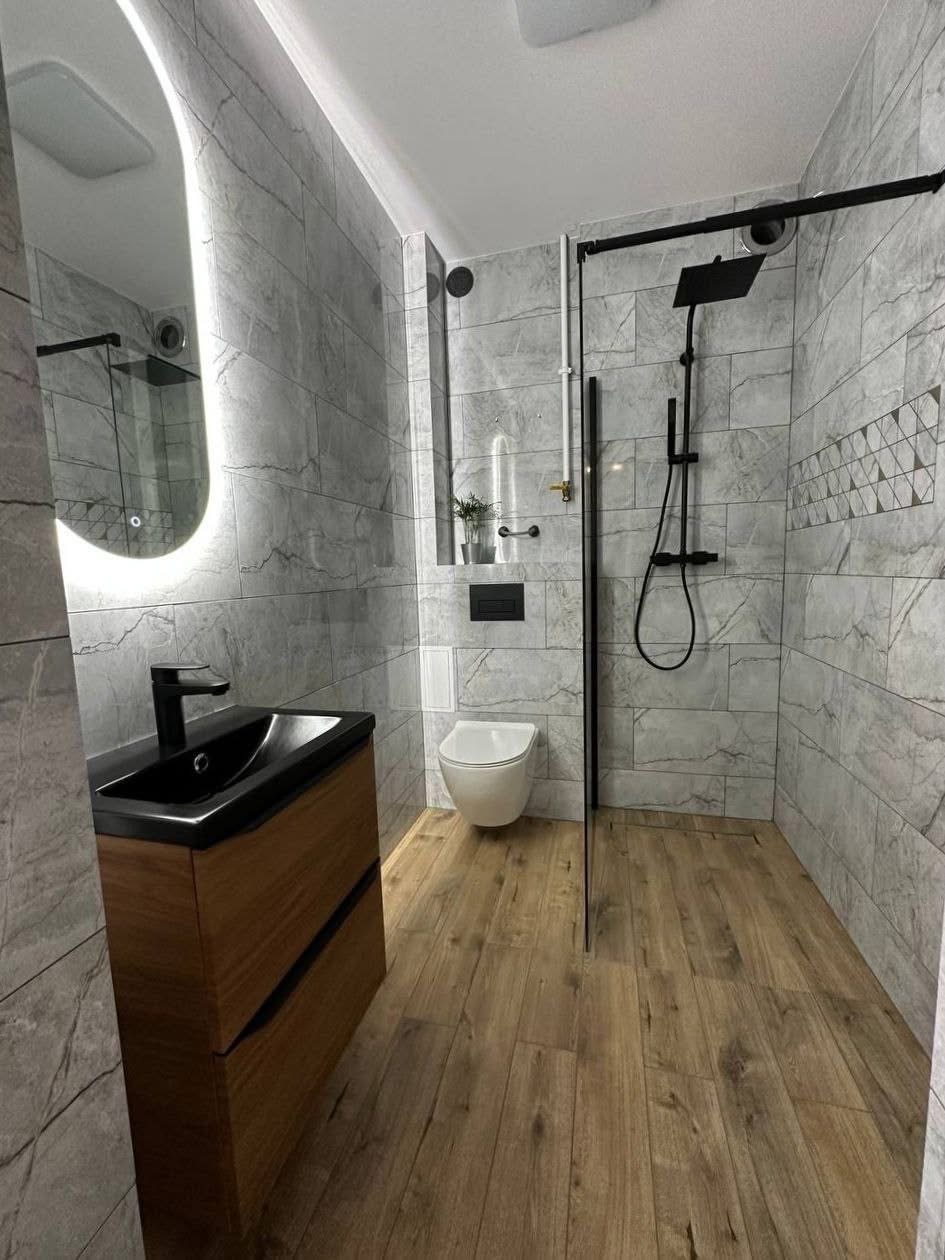 Nowoczesna łazienka z szarymi płytkami imitującymi marmur, drewnianą podłogą i czarną armaturą. Uwagę przykuwa podświetlane lustro o nieregularnym kształcie.
