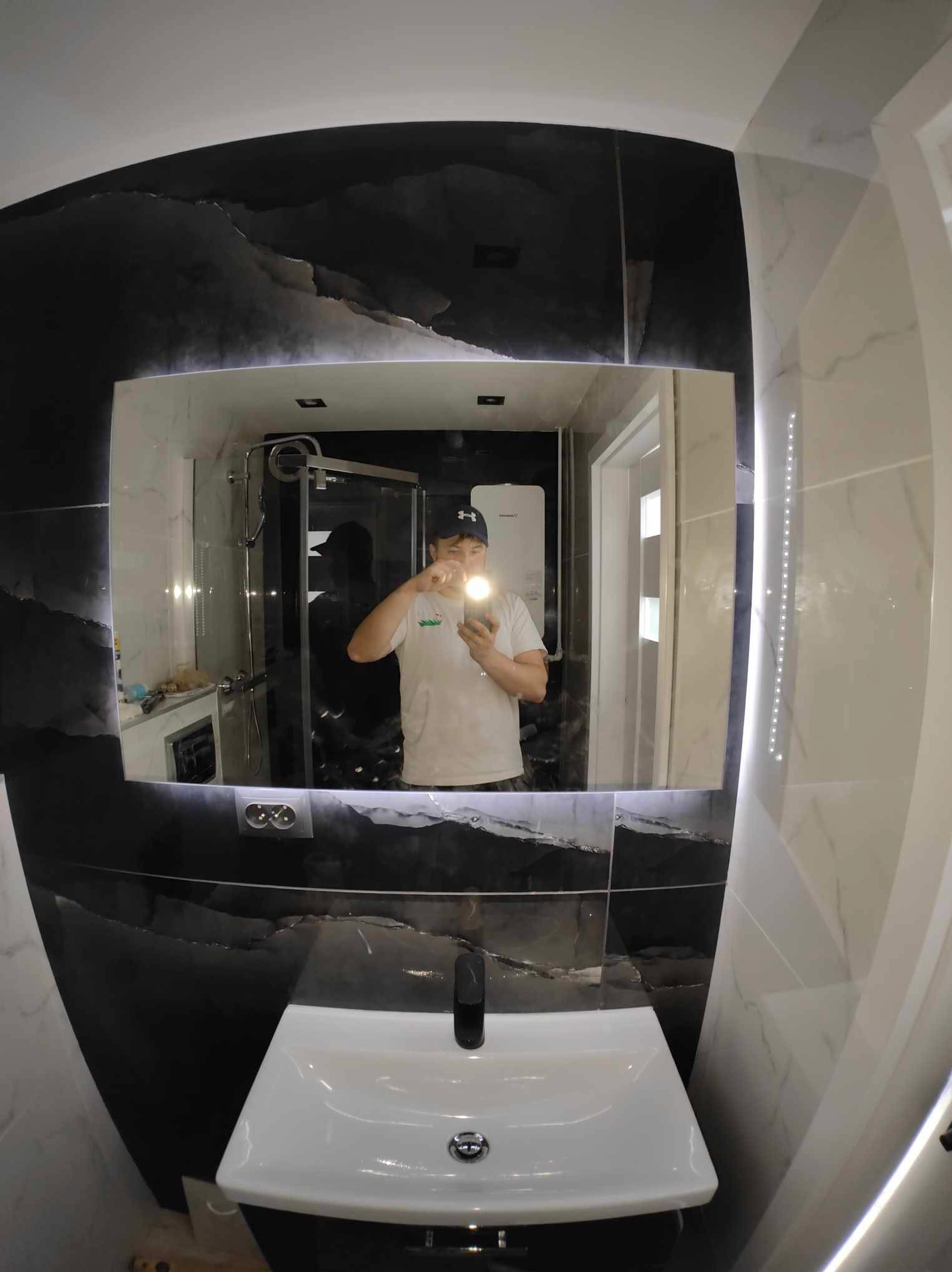 Nowoczesna łazienka z umywalką i podświetlanym lustrem, odbicie osoby robiącej zdjęcie. Czarno-białe płytki imitujące marmur na ścianach.