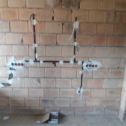 Żurowski Instalacje - Instalacja elektryczna w trakcie montażu na ceglanej ścianie, widoczne kable w peszlach i puszki podtynkowe. Na podłodze karton z narzędziami.