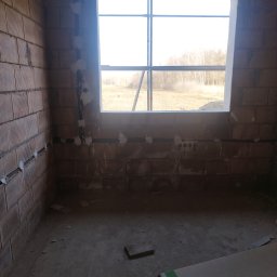 Żurowski Instalacje - Surowy stan wnętrza z instalacją elektryczną w bruzdach, widok przez okno na zewnątrz. Instalacja zabezpieczona gipsem. Ściany z czerwonej cegły.