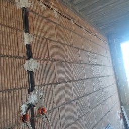 Żurowski Instalacje - Ściana z cegły z wyżłobieniami wypełnionymi przewodami elektrycznymi i puszkami instalacyjnymi. Widoczne kable. Surowy stan, w trakcie prac instalacyjnych.