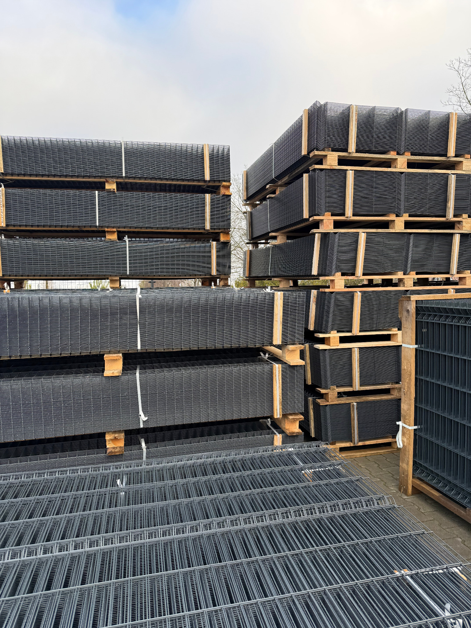 Stosy paneli ogrodzeniowych w kolorze grafitowym, ułożone na drewnianych paletach. Widoczne różne rodzaje paneli, w tym zgrzewane i ocynkowane.