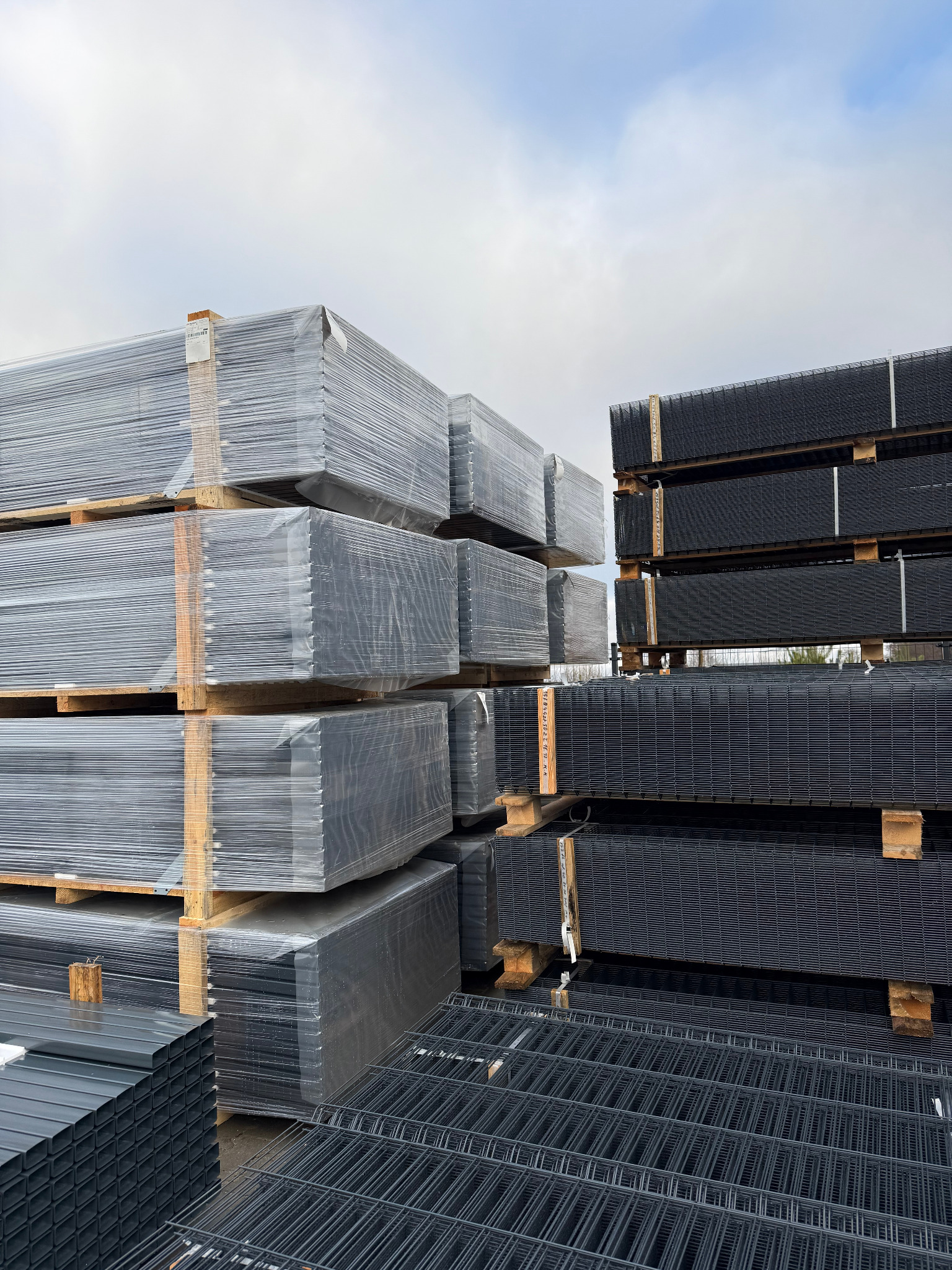 Stosy paneli ogrodzeniowych: szare, owinięte folią na paletach i czarne, metalowe panele siatkowe, gotowe do transportu i montażu. Widok z bliska.