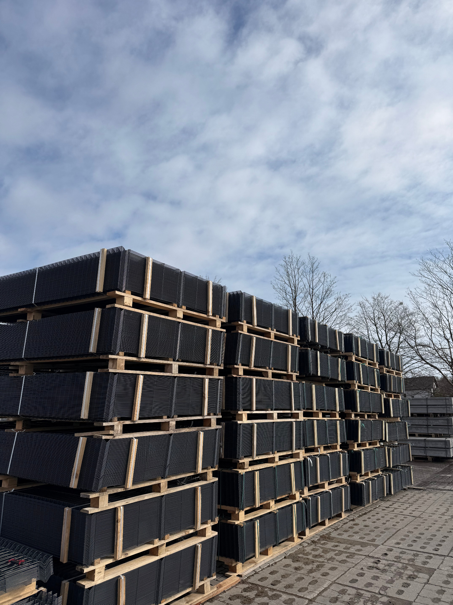 Stosy czarnych paneli ogrodzeniowych na drewnianych paletach, składowane na zewnątrz pod błękitnym niebem z chmurami. Widoczne drzewa w tle.