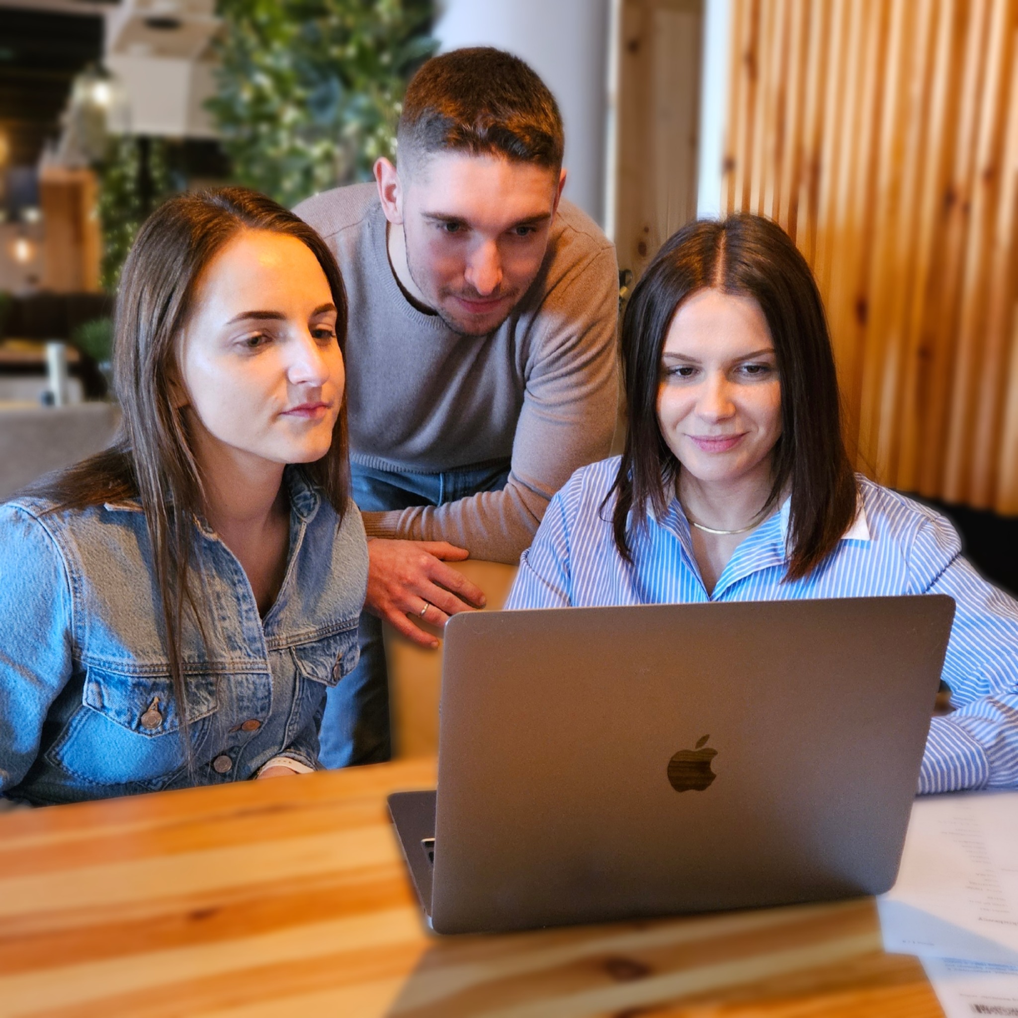 Trzy osoby pochylają się nad laptopem na drewnianym stole w jasnym wnętrzu. Dwie kobiety i mężczyzna analizują dane na ekranie. Widoczny fragment dokumentu.