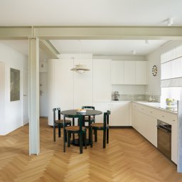 Anna Komorowicz Studio - Jasne wnętrze mieszkania z parkietem w jodełkę, okrągłym stołem i kuchnią w zabudowie. Widoczne stalowe belki konstrukcyjne. Minimalistyczny design z akcentami retro.
