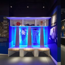 Anna Komorowicz Studio - Projekt wnętrza muzeum Lecha Poznań: szatnia z podświetlonymi koszulkami, piłką, butami i rękawicami bramkarskimi. W tle trofea w gablotach i grafiki.