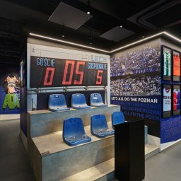 Anna Komorowicz Studio - Wnętrze muzeum piłkarskiego: trybuna z niebieskimi krzesełkami, tablica wyników, ściana z wizerunkiem kibiców i gabloty z koszulkami. Nowoczesny design, stonowana kolorystyka.