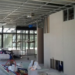 Janusz Goluch - Montaż płyt gipsowo-kartonowych w Legnicy. Widoczne profile sufitowe i ściany działowe w trakcie budowy. Na podłodze narzędzia i materiały budowlane.