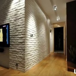 Janusz Goluch - Ściana z dekoracyjnej białej cegły w salonie z drewnianą podłogą i telewizorem. Oświetlenie punktowe podkreśla fakturę cegły.