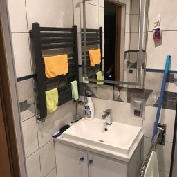 Water Service Łukasz Worek - Nowoczesna łazienka z grzejnikiem drabinkowym, umywalką na szafce i lustrem z oświetleniem. Widoczne płytki w odcieniach bieli i szarości oraz akcesoria łazienkowe.