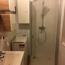 Water Service Łukasz Worek - Nowoczesna łazienka z kabiną prysznicową, umywalką i pralką. Na ścianie wiszą dwa białe grzejniki elektryczne. Widoczne płytki i armatura.