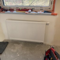 Water Service Łukasz Worek - Biały grzejnik panelowy zamontowany pod oknem, widoczne narzędzia instalacyjne na parapecie i torba narzędziowa Milwaukee na podłodze. Świeżo zamontowany grzejnik.