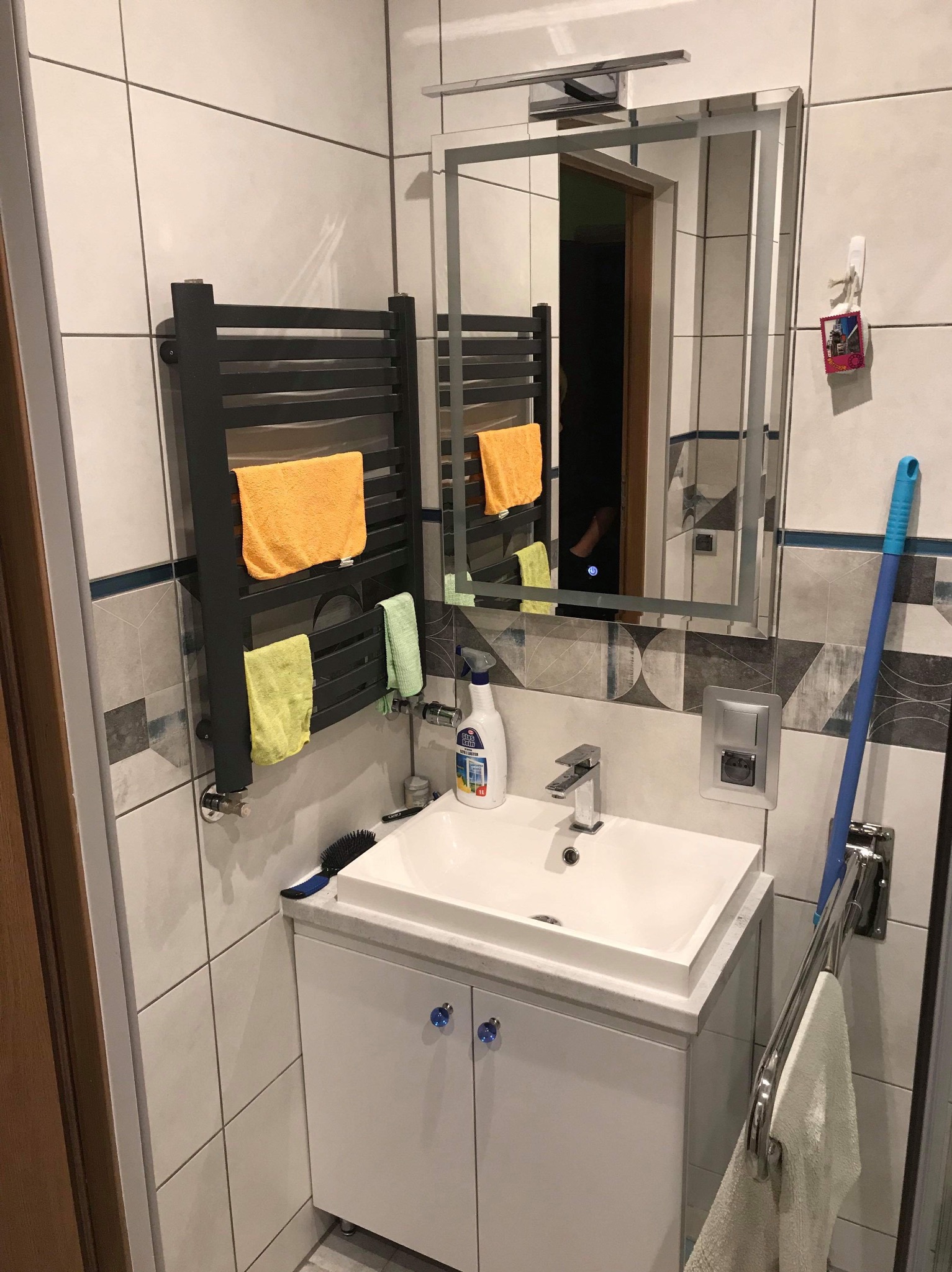 Nowoczesna łazienka z grzejnikiem drabinkowym, umywalką na szafce i lustrem z oświetleniem. Widoczne płytki w odcieniach bieli i szarości oraz akcesoria łazienkowe.