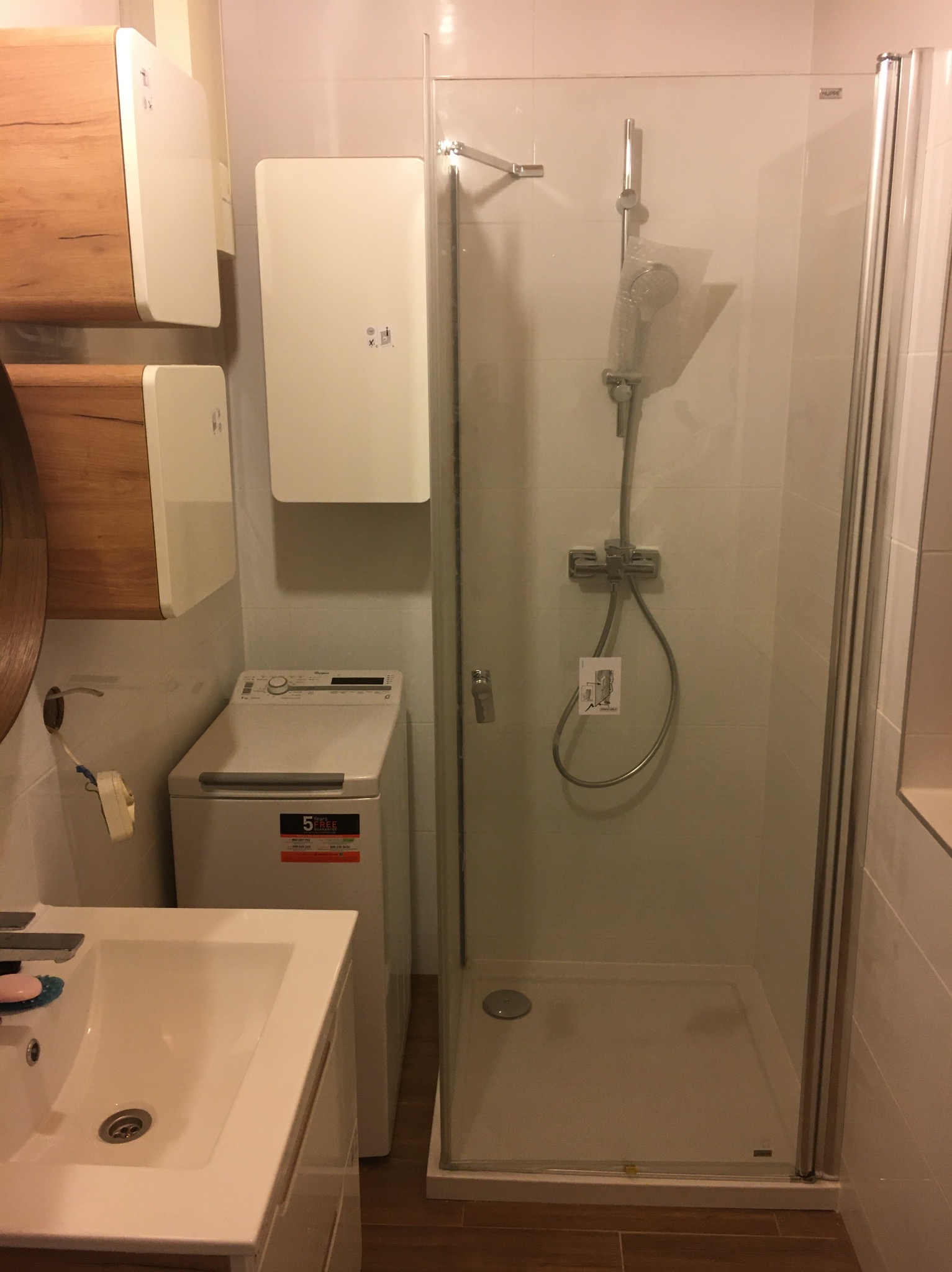 Nowoczesna łazienka z kabiną prysznicową, umywalką i pralką. Na ścianie wiszą dwa białe grzejniki elektryczne. Widoczne płytki i armatura.