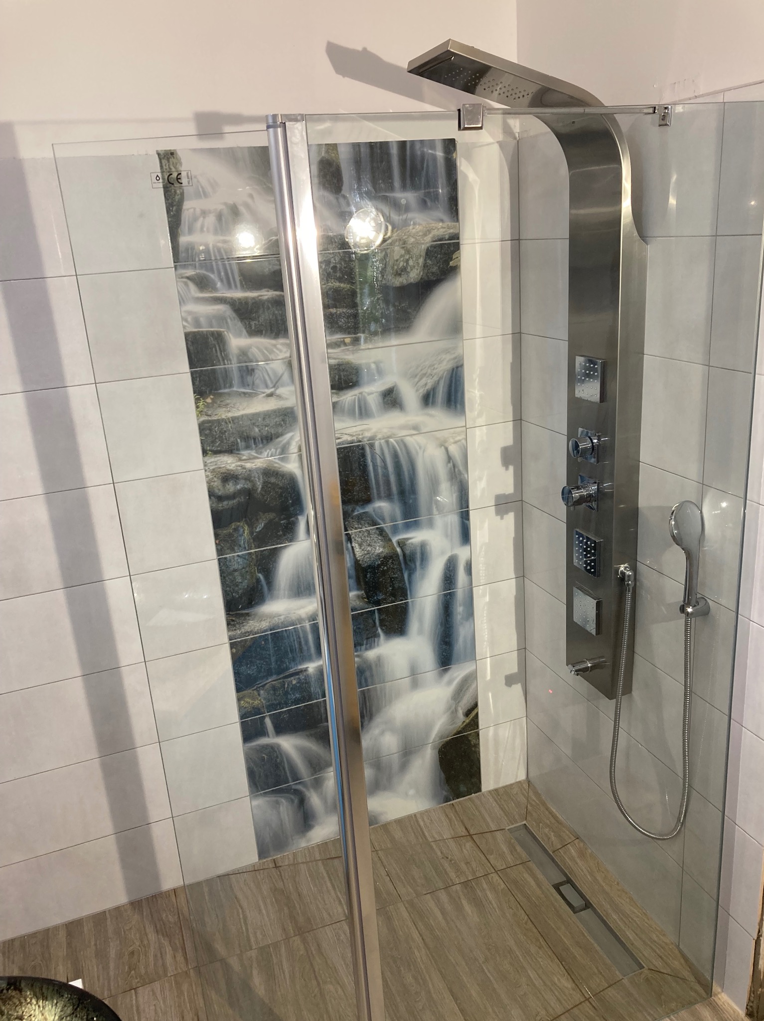 Nowoczesna kabina prysznicowa z panelem natryskowym i płytkami z motywem wodospadu. Szklane drzwi z chromowanymi okuciami. Wykończenie w jasnych, neutralnych barwach.