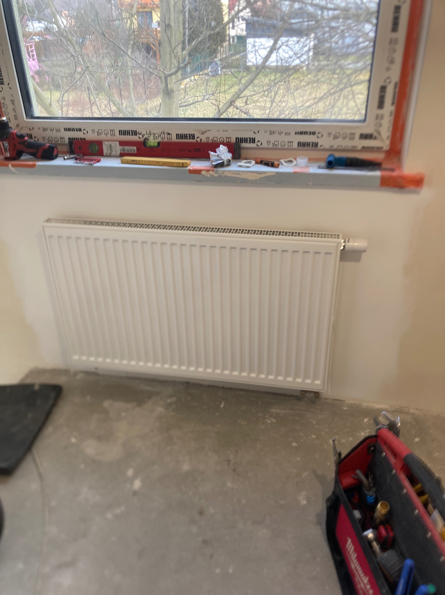 Biały grzejnik panelowy zamontowany pod oknem, widoczne narzędzia instalacyjne na parapecie i torba narzędziowa Milwaukee na podłodze. Świeżo zamontowany grzejnik.