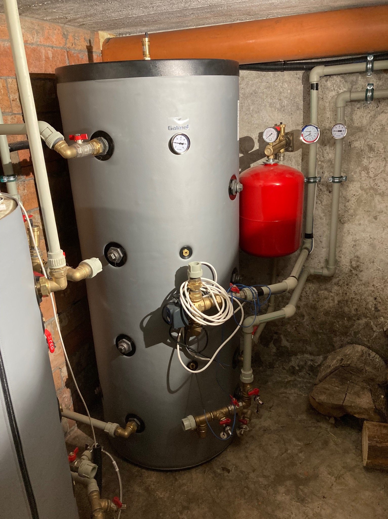 Szary bufor ciepła Galmet z czerwoną przeponą, manometrami i rurami w piwnicy z ceglaną ścianą. Widoczne liczne zawory i przyłącza.