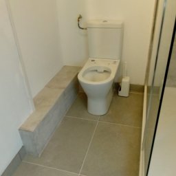 Łukasz - Wykończona łazienka z toaletą na podwyższeniu, jasne płytki podłogowe i częściowo widoczna kabina prysznicowa. Minimalistyczny design.