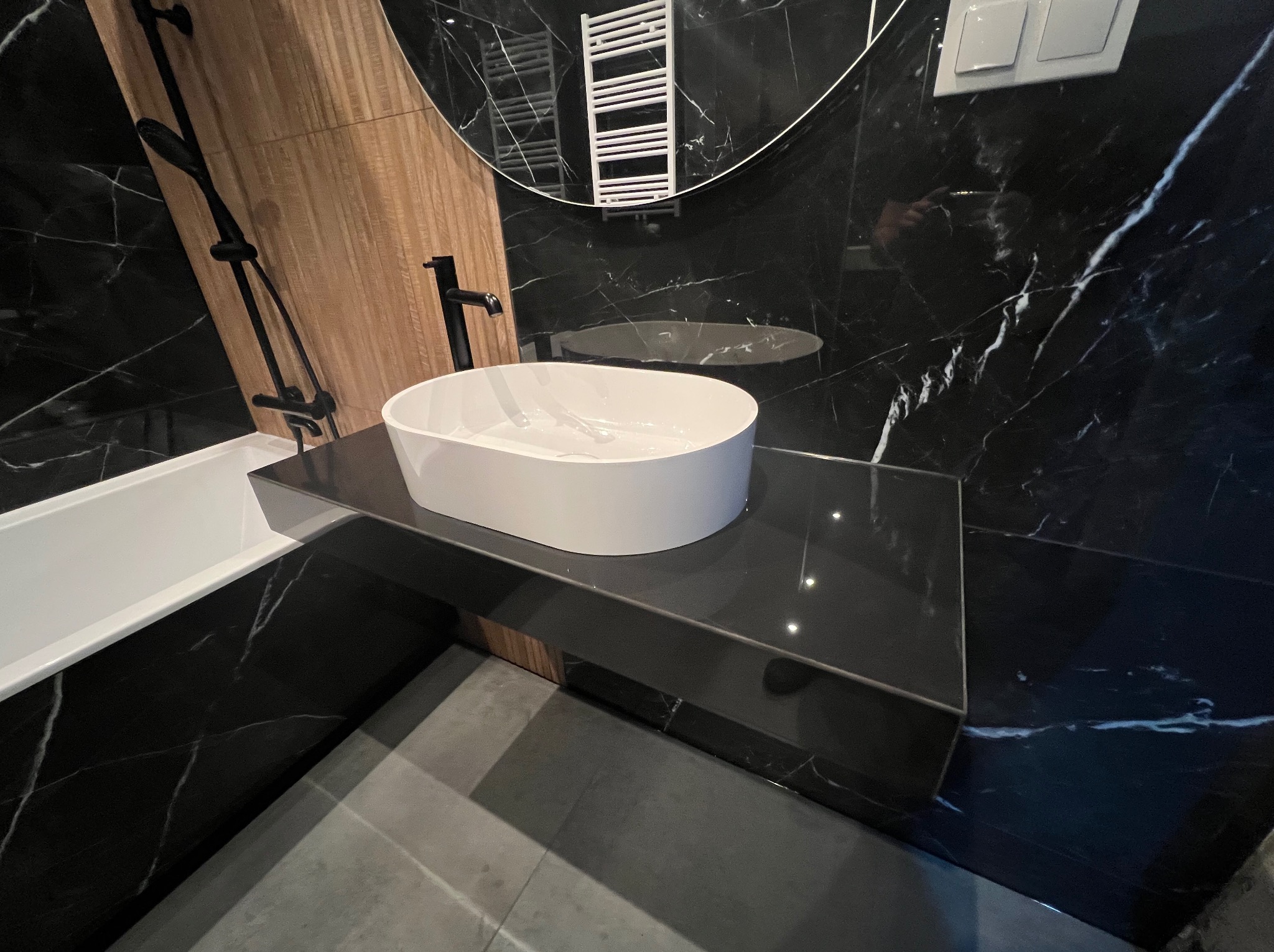 Nowoczesna łazienka z czarnymi marmurowymi płytkami, owalną białą umywalką na czarnym blacie i elementami drewna. Widoczne bateria, grzejnik i wanna.