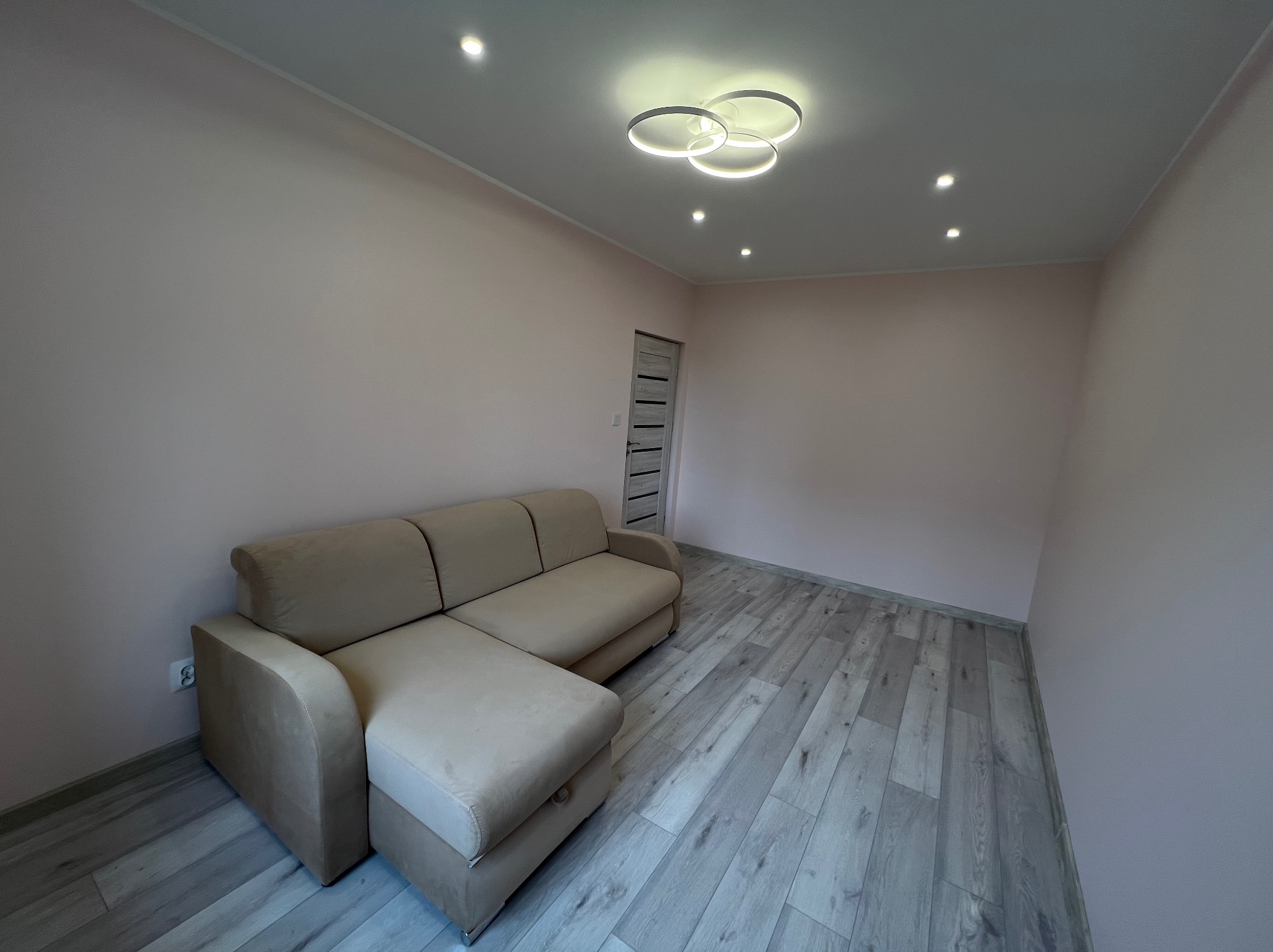 Jasny salon po remoncie: beżowa sofa narożna, panele podłogowe imitujące drewno, ściany w odcieniach różu, nowoczesna lampa sufitowa z trzema okręgami.