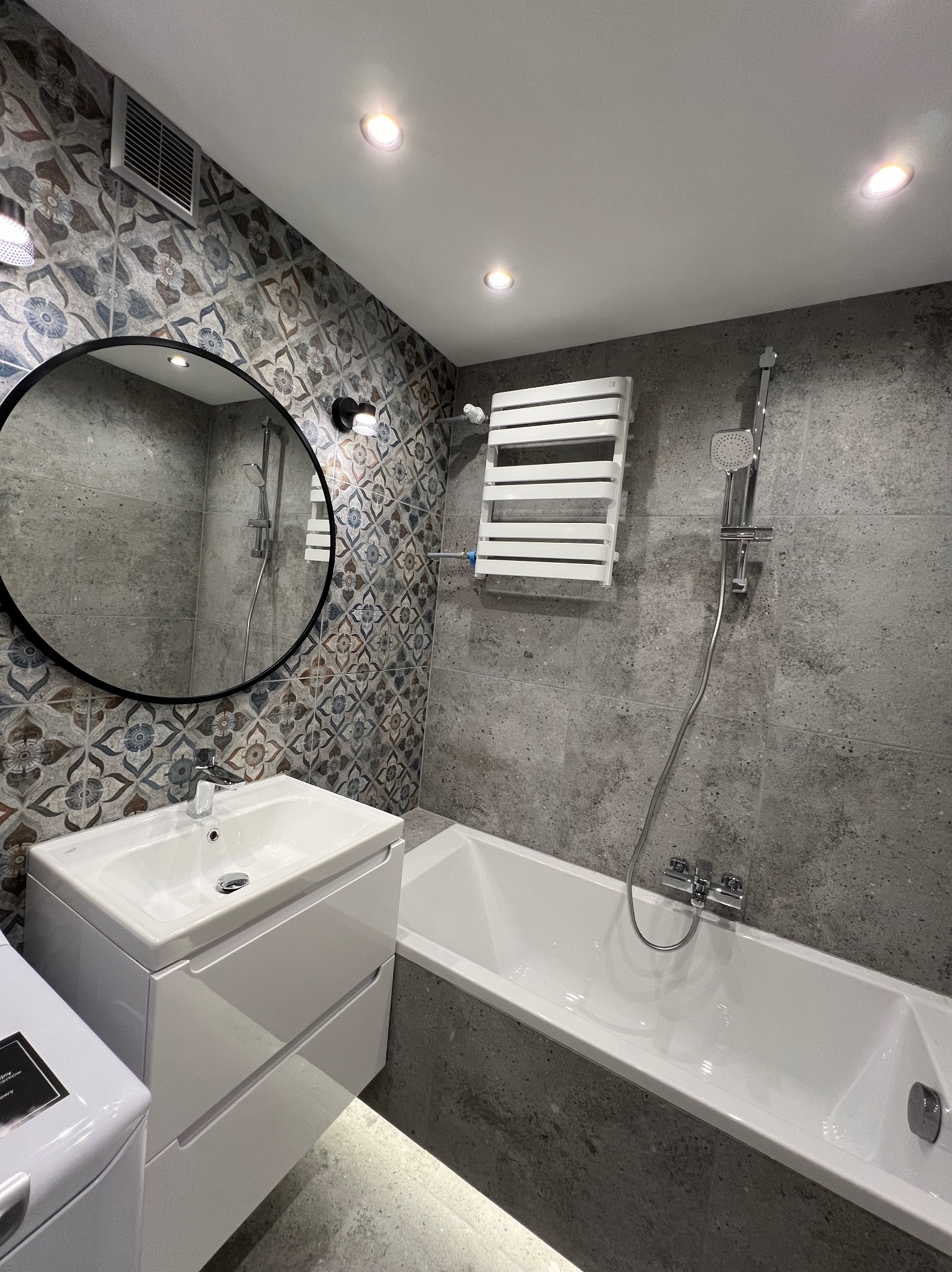 Nowoczesna łazienka z wanną, umywalką z szafką i okrągłym lustrem. Ściany wyłożone płytkami w odcieniach szarości i wzorzystymi płytkami dekoracyjnymi.