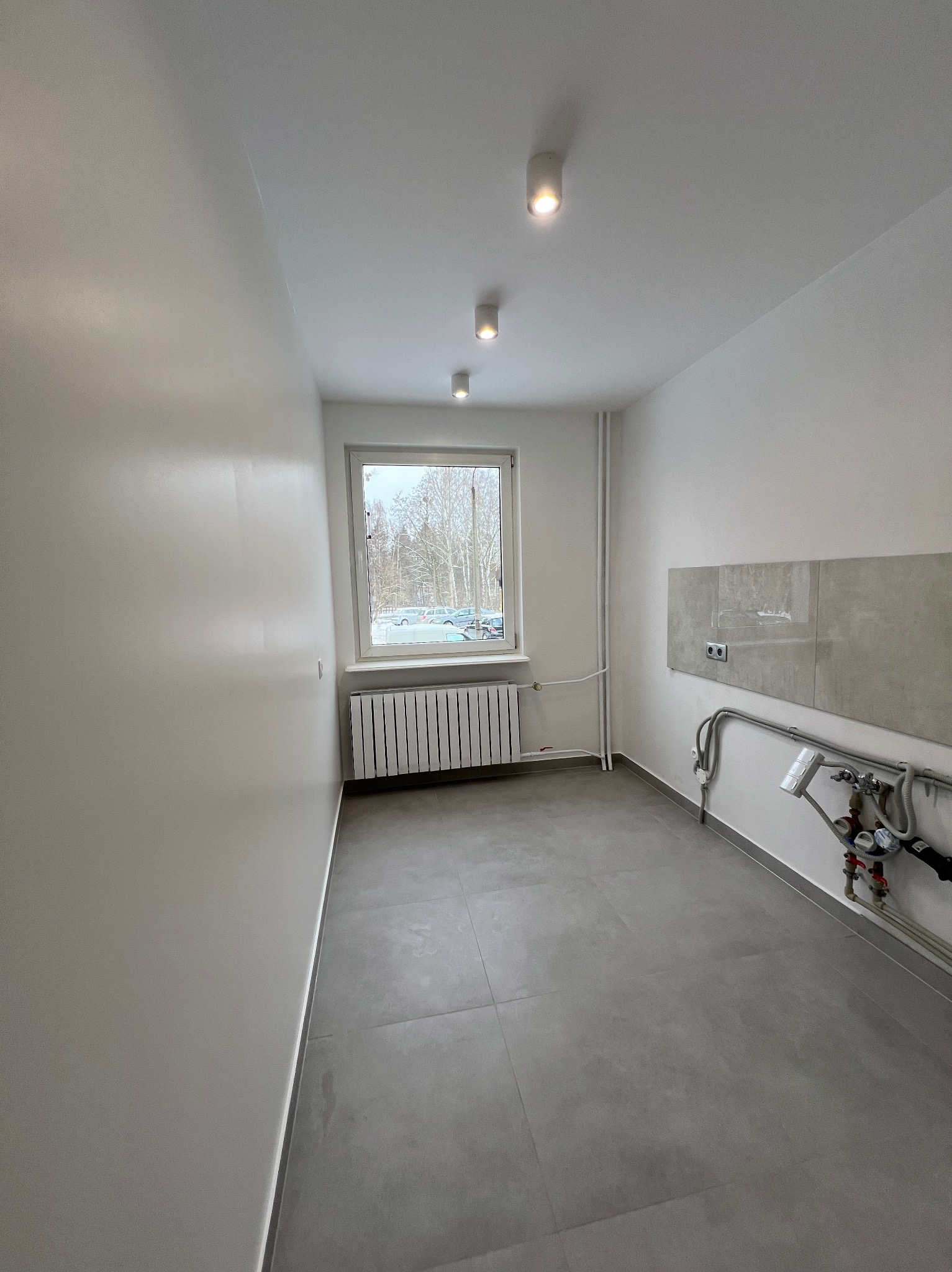 Wykończone wnętrze łazienki z szarymi płytkami podłogowymi i częściowo na ścianie, białe ściany, grzejnik pod oknem, instalacja wodna przy ścianie, trzy lampy sufitowe.