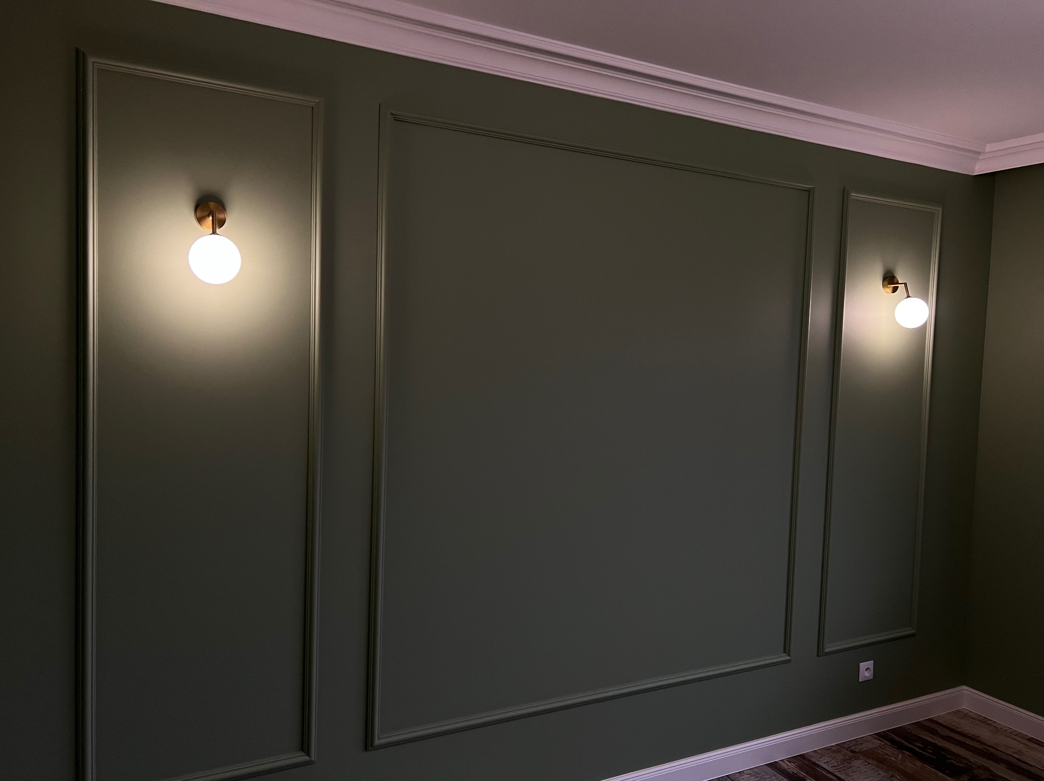 Elegancka ściana w odcieniach zieleni z dekoracyjnymi listwami i dwoma okrągłymi lampami ściennymi, podkreślającymi klasyczny charakter wnętrza.