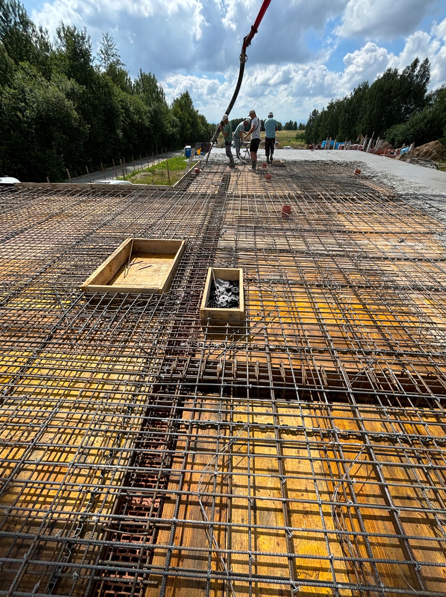 Zbrojenie stropu z deskowaniem i betonowaniem w tle. Widoczni pracownicy i pompa do betonu. Dwa drewniane elementy na pierwszym planie. Prace budowlane w toku.