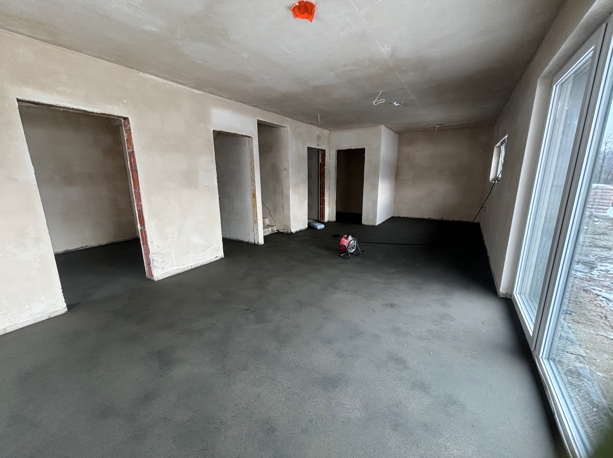 Wnętrze w stanie deweloperskim po wylaniu posadzki. Widoczne surowe ściany, otwory drzwiowe i okno. Na podłodze stoi czerwona nagrzewnica.