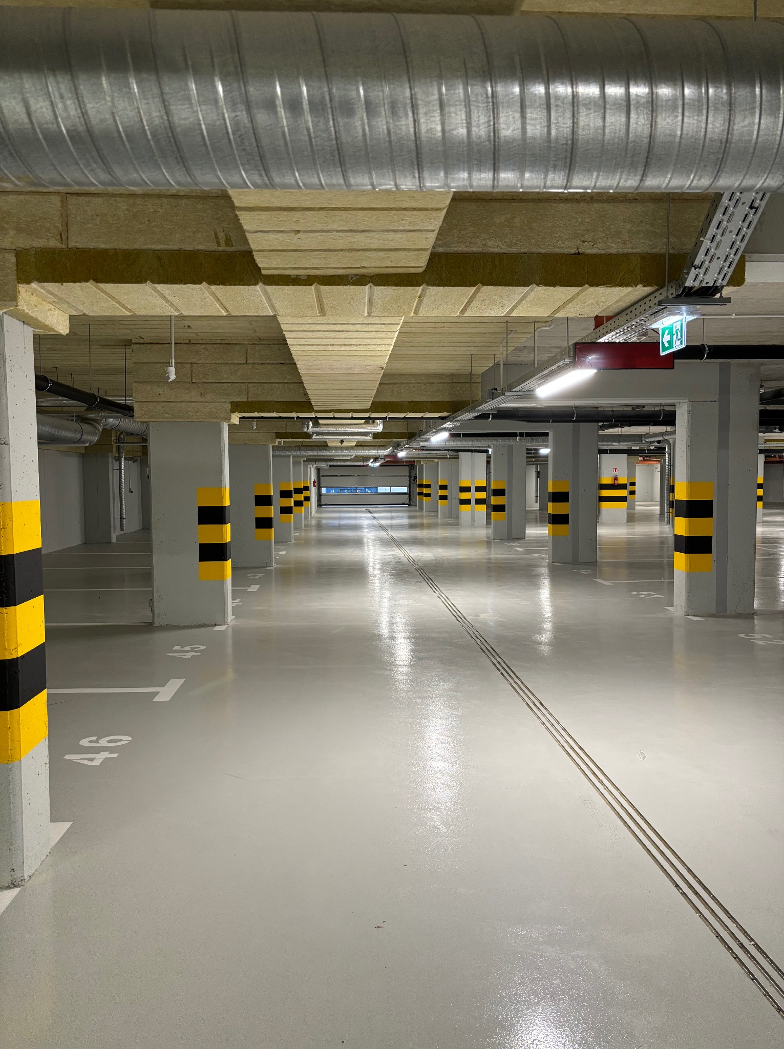 Pusty parking podziemny z betonowymi kolumnami w żółto-czarne pasy, widoczne instalacje pod sufitem i numery miejsc parkingowych na szarej posadzce.