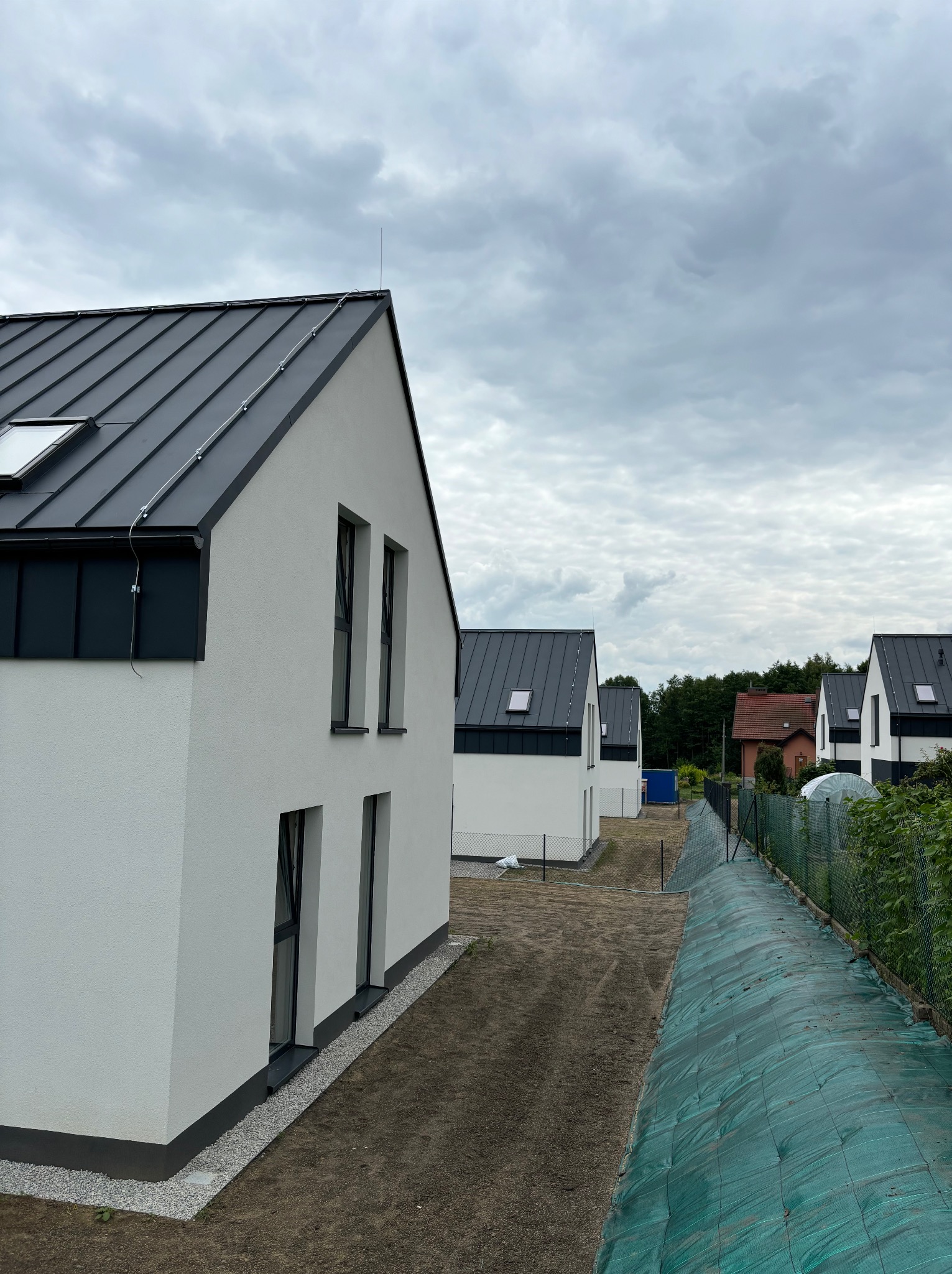 Nowoczesne, białe domy jednorodzinne z ciemnymi dachami i oknami, w trakcie budowy, widok z góry na osiedle w pochmurny dzień. Widoczny fragment ogrodzenia.