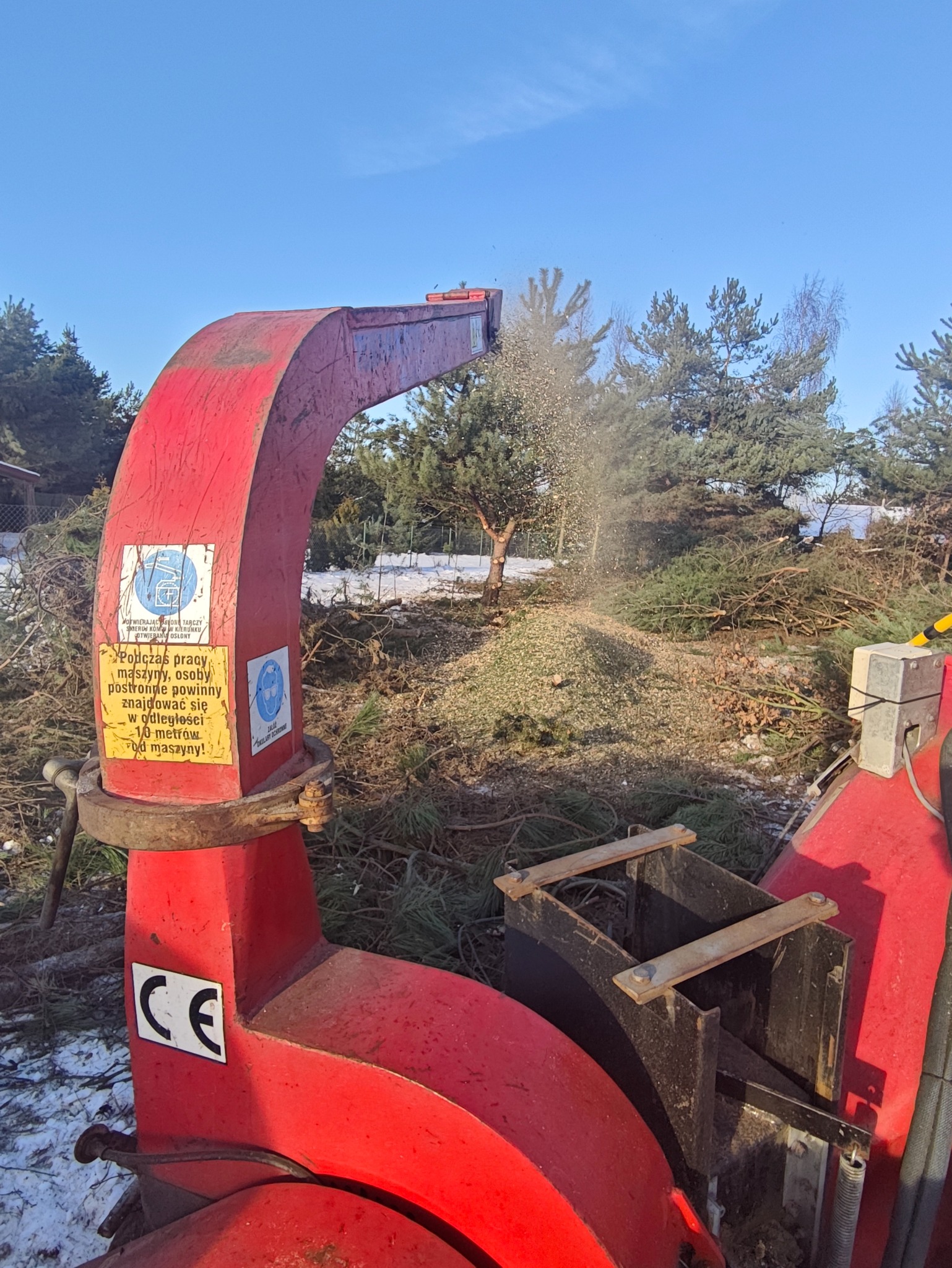 Czerwony rębak do gałęzi w akcji, w tle stosy pociętych gałęzi i drzewa. Wokół resztki śniegu. Widoczne ostrzeżenia na maszynie.
