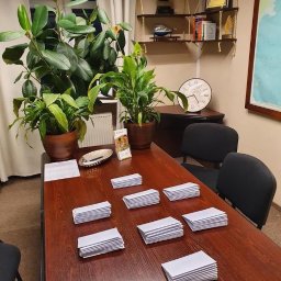 KANCELARIA FINANSOWA CENTRUM ODDŁUŻEŃ SPÓŁKA Z OGRANICZONĄ ODPOWIEDZIALNOŚCIĄ - Biuro z drewnianym stołem, na którym leżą pliki białych kopert. W tle rośliny doniczkowe, półki z dekoracjami i mapa świata. Przytulne wnętrze kancelarii.