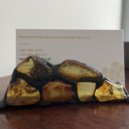 KANCELARIA FINANSOWA CENTRUM ODDŁUŻEŃ SPÓŁKA Z OGRANICZONĄ ODPOWIEDZIALNOŚCIĄ - Wizytówka Kancelarii Finansowej Centrum Oddłużeń umieszczona w unikalnym, bursztynowym stojaku na drewnianym biurku. Widoczne dane kontaktowe firmy.